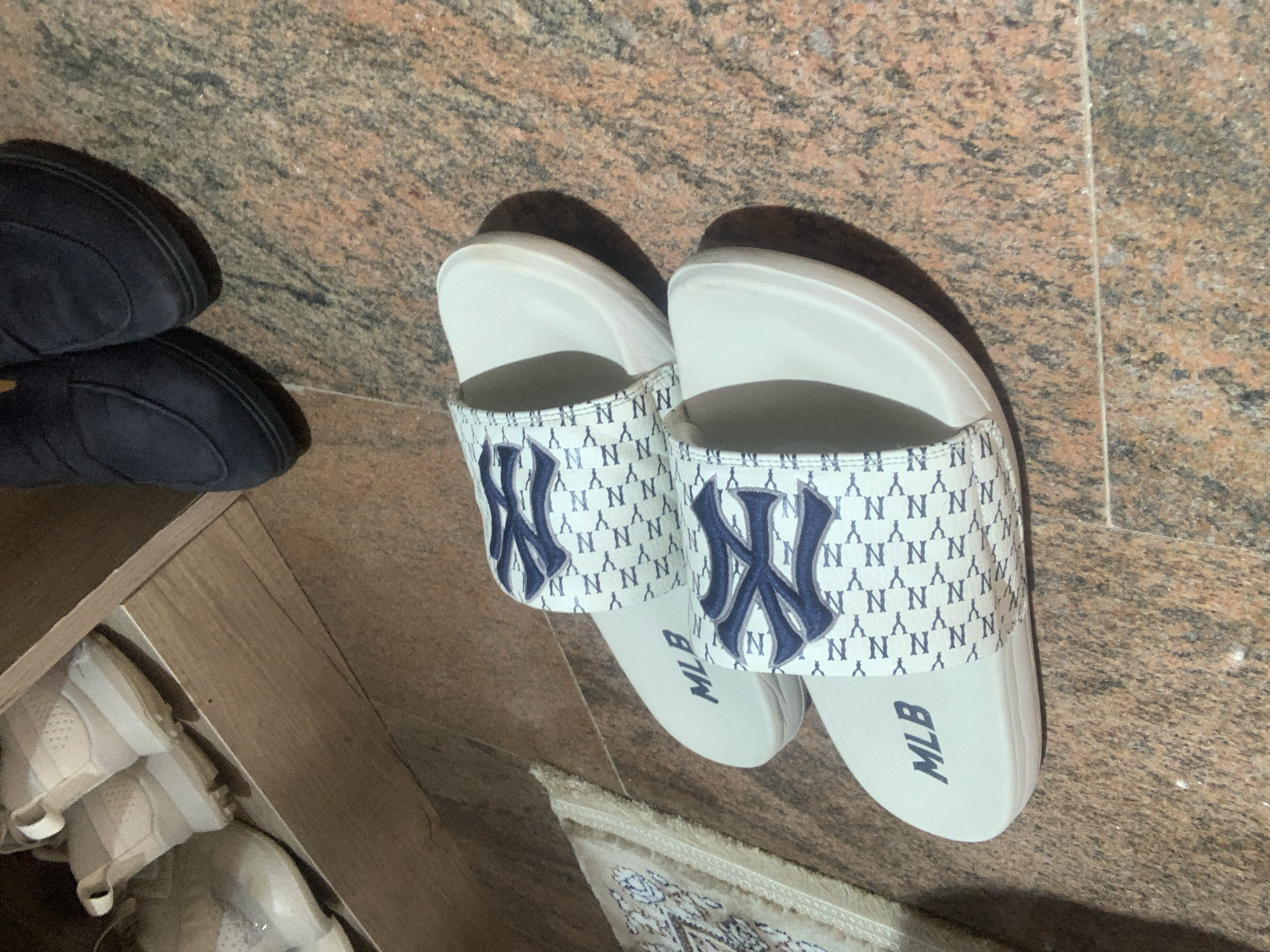 Two white MLB slides with a navy blue New York Yankees logo pattern are displayed on a speckled brown and pink tiled floor. Next to the slides are two dark navy blue slip-on shoes. In the background, a wooden shelf contains several pairs of light-colored sneakers. The scene is likely from a residential setting in Abuja, Nigeria.