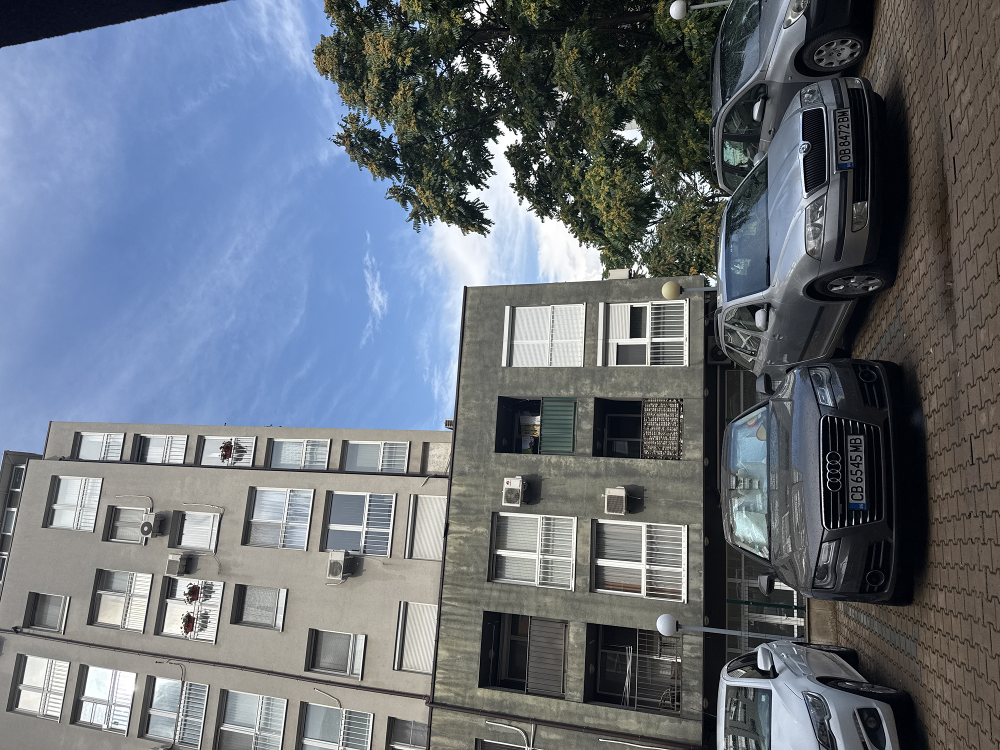 The image shows a parking lot in front of apartment buildings in Sofia, Bulgaria. Several cars are parked in the lot, including a dark gray Audi, a silver Skoda station wagon, and a white car. The apartment buildings are tall and gray, with many windows and balconies. Trees with green and yellow leaves are visible in the background. The sky is blue with some white clouds. The ground in the parking lot is paved with red bricks.
