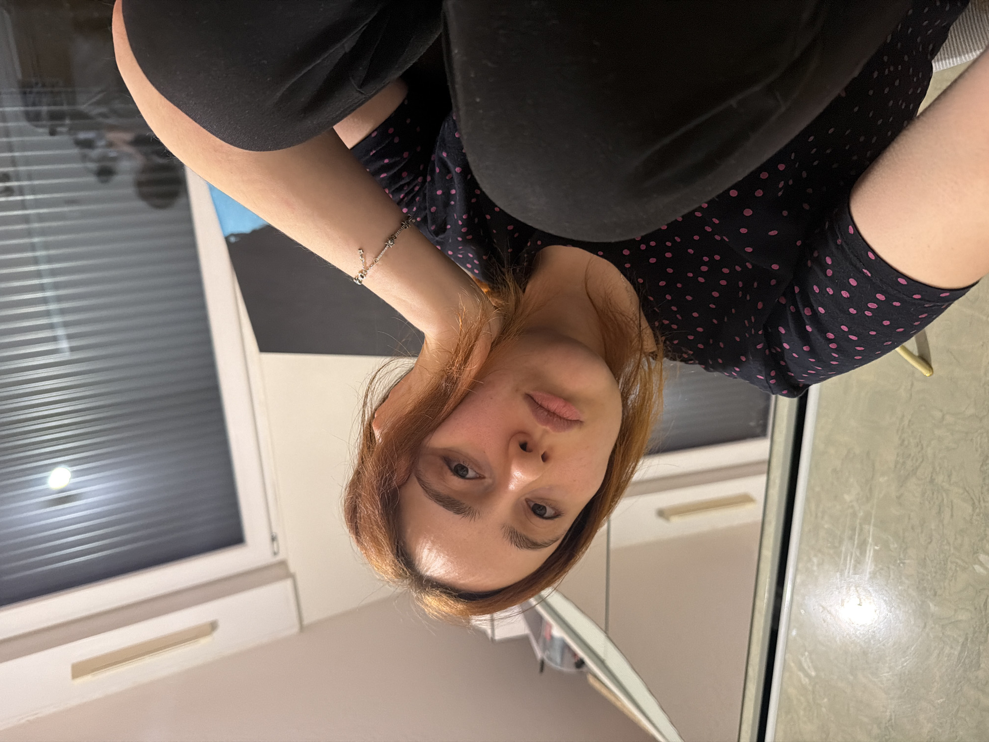 A woman is posing for a selfie in what appears to be a kitchen or bathroom with neutral-toned cabinets and a window featuring closed blinds. She is wearing dark clothing, with a top that has a polka dot pattern, and dark pants. The woman is wearing a bracelet on her left wrist. The lighting in the room is dim. The location information is not evident in the image to indicate the specific city in France.