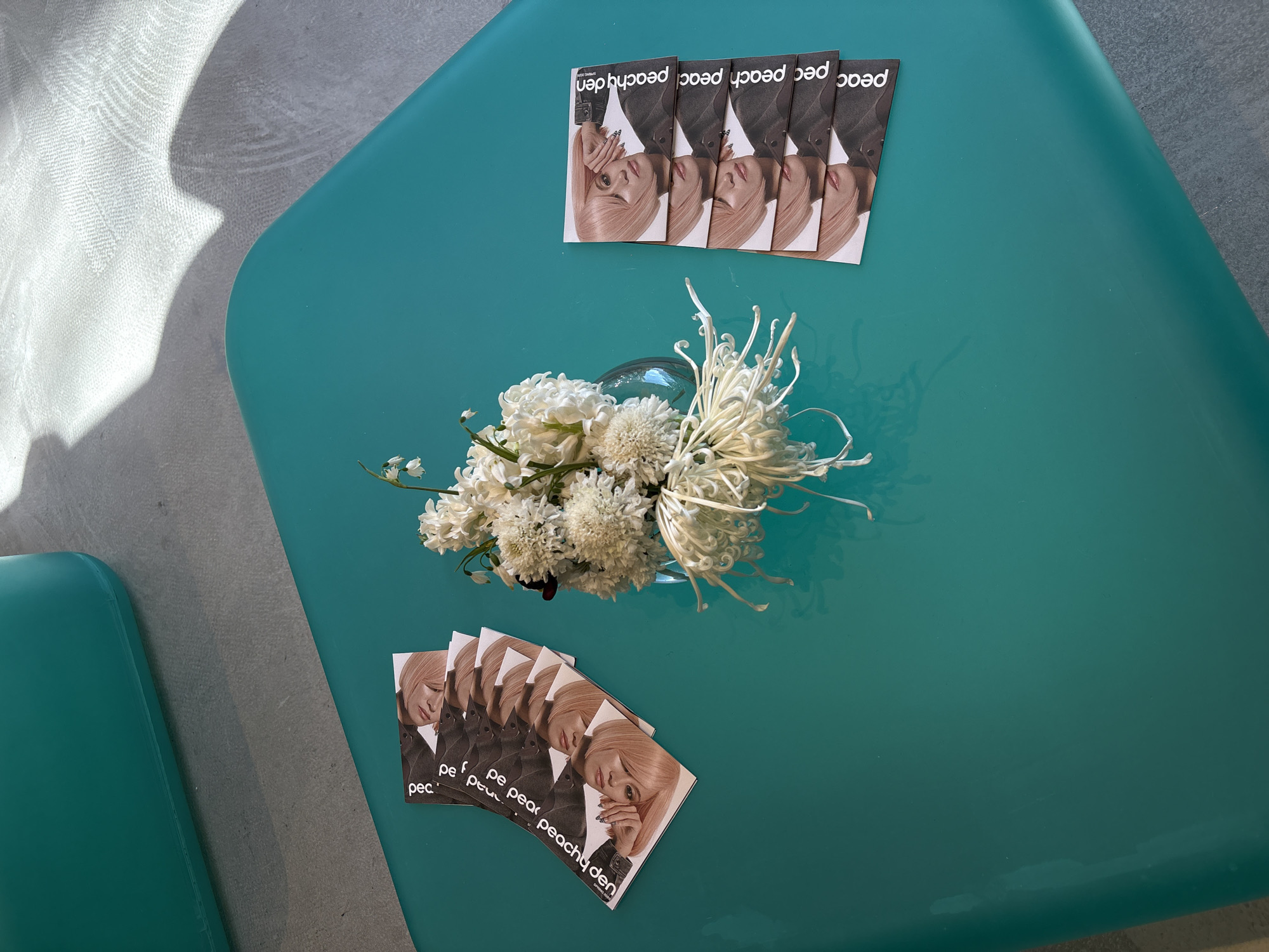 The image shows an overhead view of a bright turquoise surface, possibly a table or seat, with two clusters of identical brochures on it. Each brochure features a portrait of a fair-skinned person with blonde hair. The name "peachy den" is printed on the brochures. A small clear vase filled with white flowers sits in the center of the surface, between the two groups of brochures. Part of a matching turquoise seat or surface is visible on the left side of the frame. Light falls from the upper left, casting a shadow on the textured surface in that area. The location appears to be an indoor space in the city of London, United Kingdom.