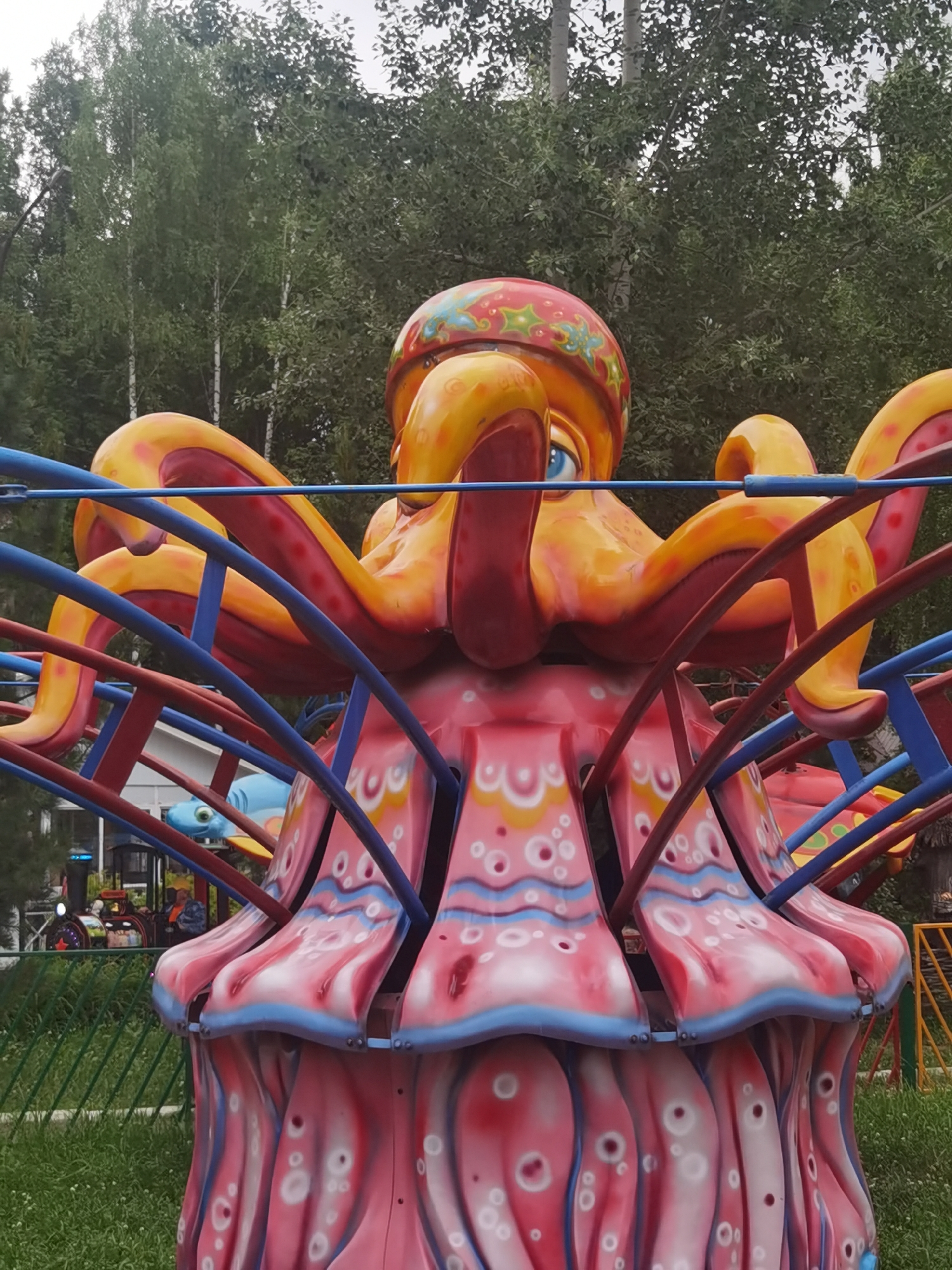 The image shows a close-up of a colorful amusement park ride designed to resemble an octopus. The ride is predominantly orange and pink, with blue and red accents from its supporting structure. The octopus head is large and cartoonish, with a prominent blue eye and decorative red and yellow patterns on its head. Its tentacles are curved and bright orange, appearing to hold up the central part of the ride. The lower section of the ride is pink with stylized white and blue markings, resembling the tentacles or a skirt. In the background, there are green trees and a cloudy sky. Partially visible to the left and behind the ride are other amusement park elements, including a small train-like ride with people inside and a green metal fence. The overall setting appears to be an outdoor amusement park, likely in Chaikovsky, Russia, given the context. The image is taken from a low angle, focusing on the detail and vibrant colors of the octopus ride.