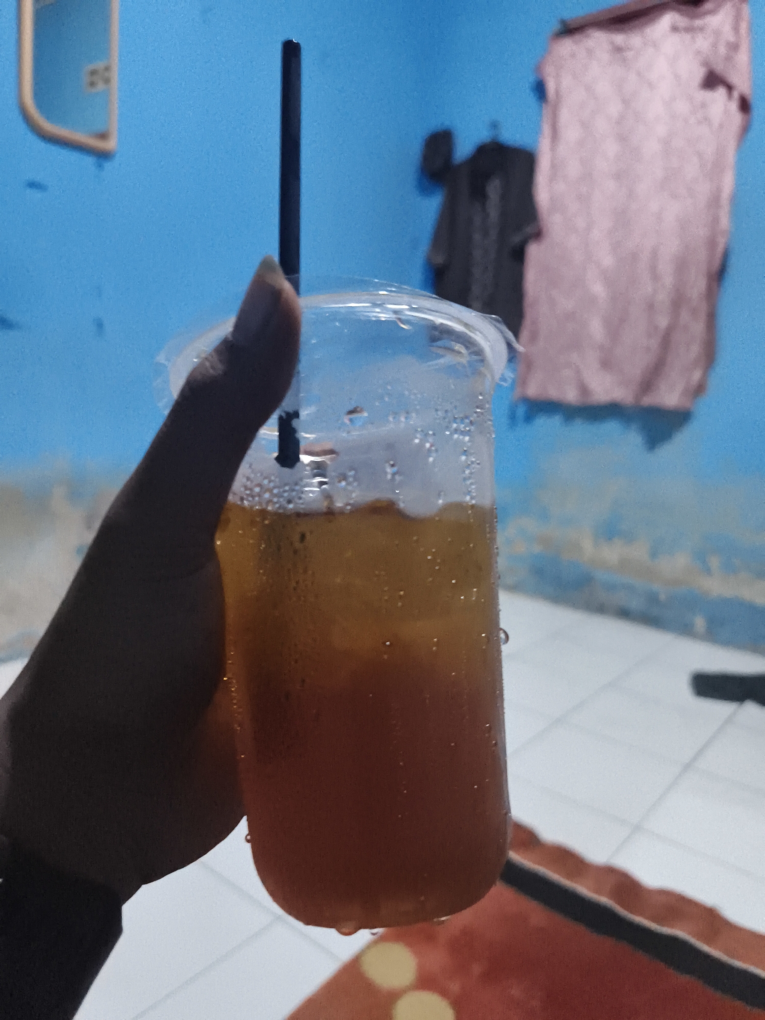 A hand holds a clear plastic cup filled with a dark orange beverage and ice. Condensation on the outside of the cup suggests it is cold. A black straw extends from the drink. The background is a bright blue wall with a pinkish-purple garment hanging on it, and a dark garment on a hanger to the left. A white tiled floor is visible below. The scene is likely an indoor domestic setting in Pangkalpinang, Indonesia, given the characteristic blue walls often found in local homes and the typical attire.