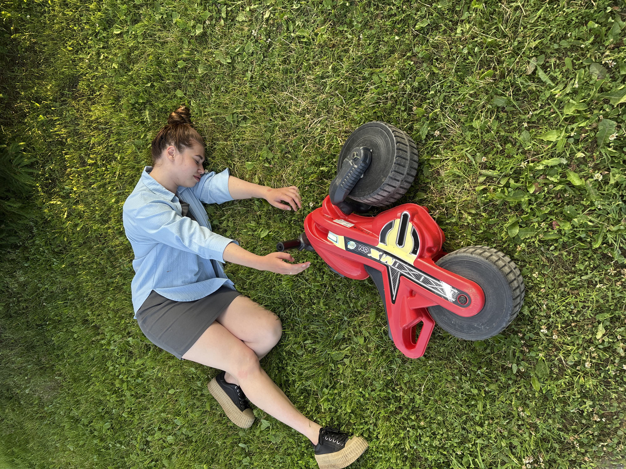 A young woman with her hair in a bun is lying on her back in the grass, appearing to be asleep. She is wearing a light blue button-up shirt and a dark gray skirt, paired with black platform shoes. Her right arm is extended towards a red toy motorcycle with black wheels, which is lying on its side next to her. The toy motorcycle has yellow and black graphics. The scene is set on a grassy lawn, likely outdoors, with dappled sunlight suggesting it is daytime. The overall mood appears peaceful and relaxed. The location context is identified as Мещёры, Russia. There is some visible text on the toy motorcycle, appearing to be "MX ON".