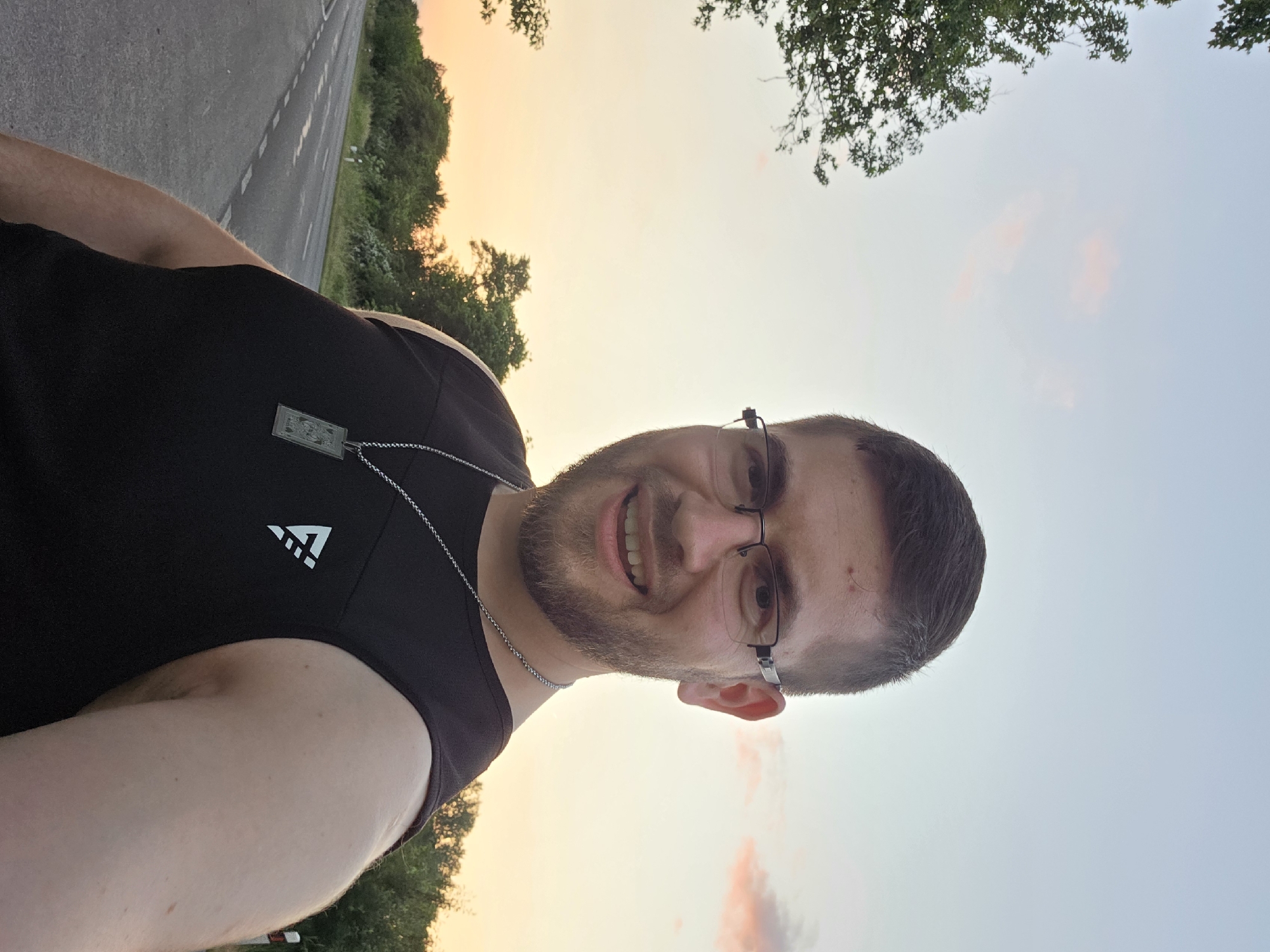 A man is depicted in a selfie style photograph. The man is wearing glasses and a black tank top with a white logo. He also wears a silver chain with a small rectangular pendant. He is smiling, and he has a beard.
The background shows a road and vegetation. The sky has a gradient of colors, from yellow to light blue, suggesting either sunrise or sunset. Based on the description, the photograph was taken outdoors in Cheshnegirovo, Bulgaria.