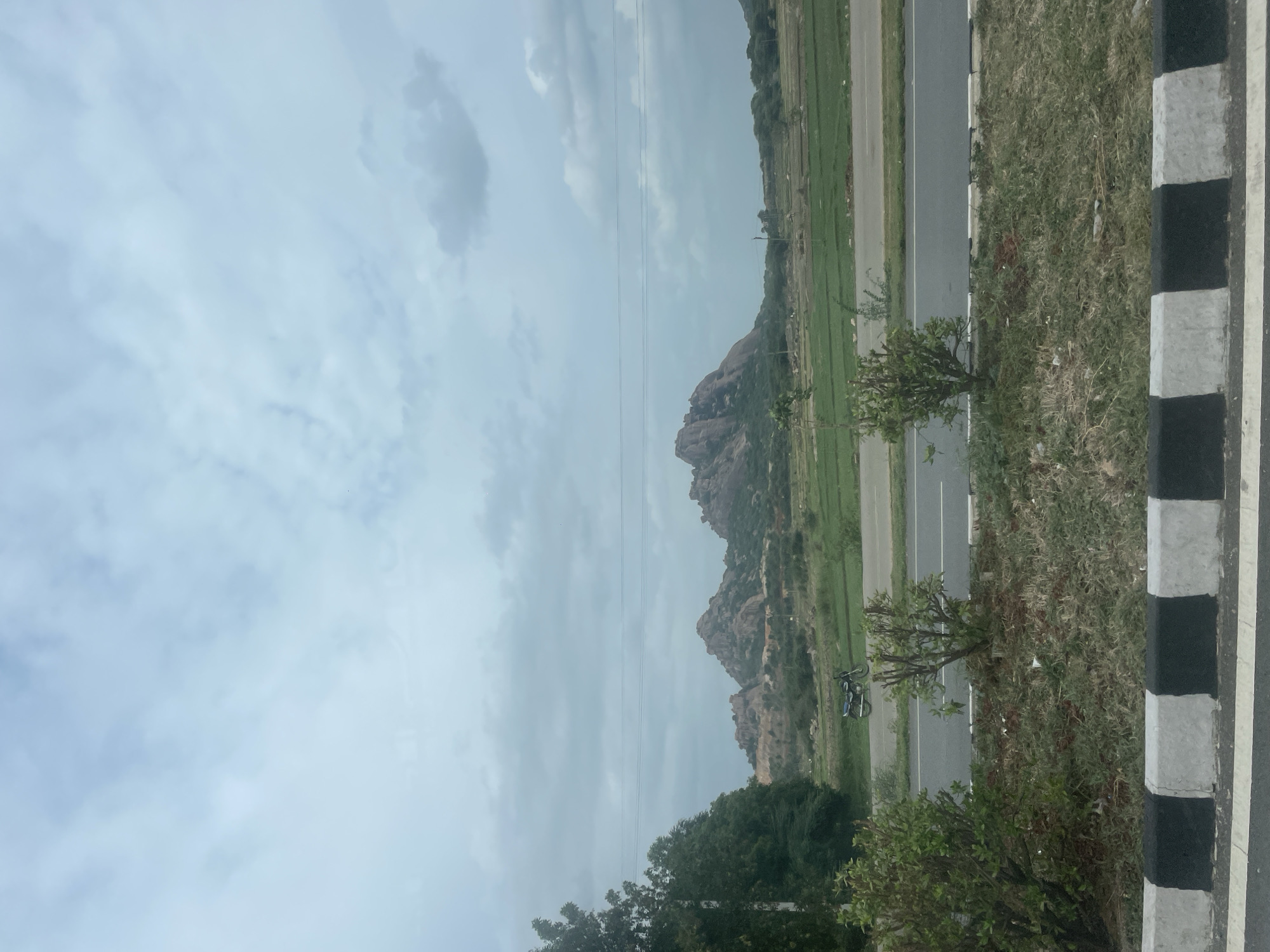A scenic view captures a road passing through a landscape with rocky hills and green fields under a cloudy sky. In the foreground, the edge of a road is visible with a black and white concrete divider. Beyond the divider, grass and some small bushes grow. A two-lane road stretches horizontally across the frame, with a motorcycle parked on the side. In the middle ground, green fields are cultivated, and trees dot the landscape. The background is dominated by a range of rugged, rocky hills. The sky is overcast with scattered clouds. The overall scene appears to be from the outskirts of Addakal, India, a region known for its natural beauty and agrarian settings.