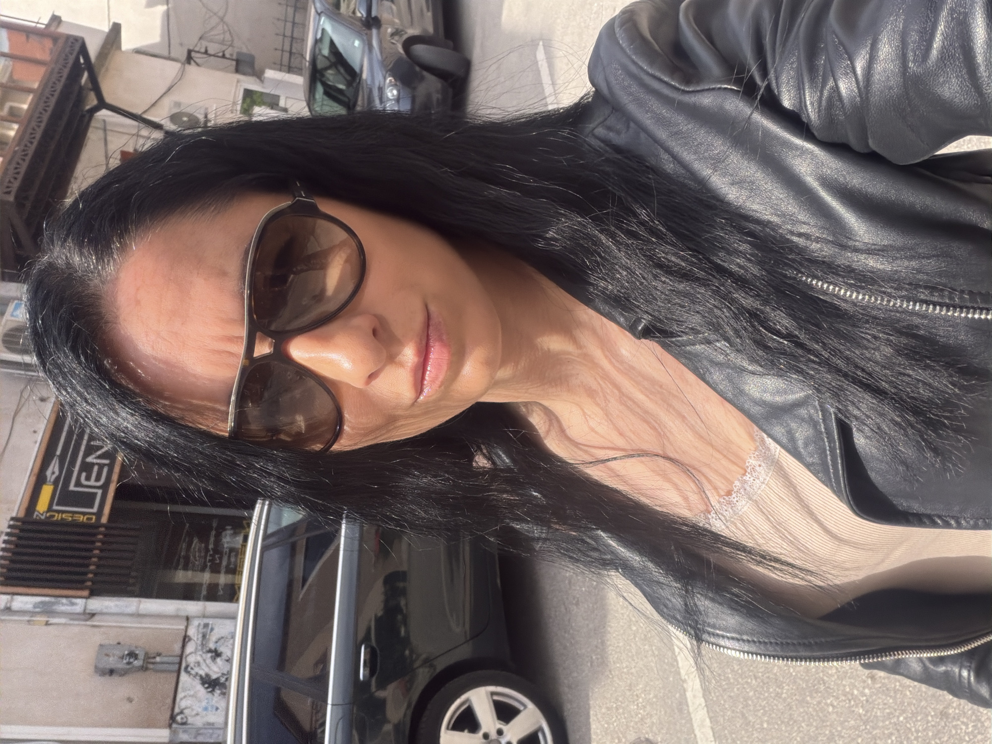 A medium-shot, canted, close-up depicts a dark-haired woman wearing sunglasses and a black leather jacket. The scene is outdoors in an urban environment. The background reveals buildings and parked vehicles, including a black car. "DESIGN" is visible on a sign behind the woman. Given the location, it is assumed the environment is in the city of Pleven, Bulgaria. The woman is in the foreground, appearing to be taking a selfie.
