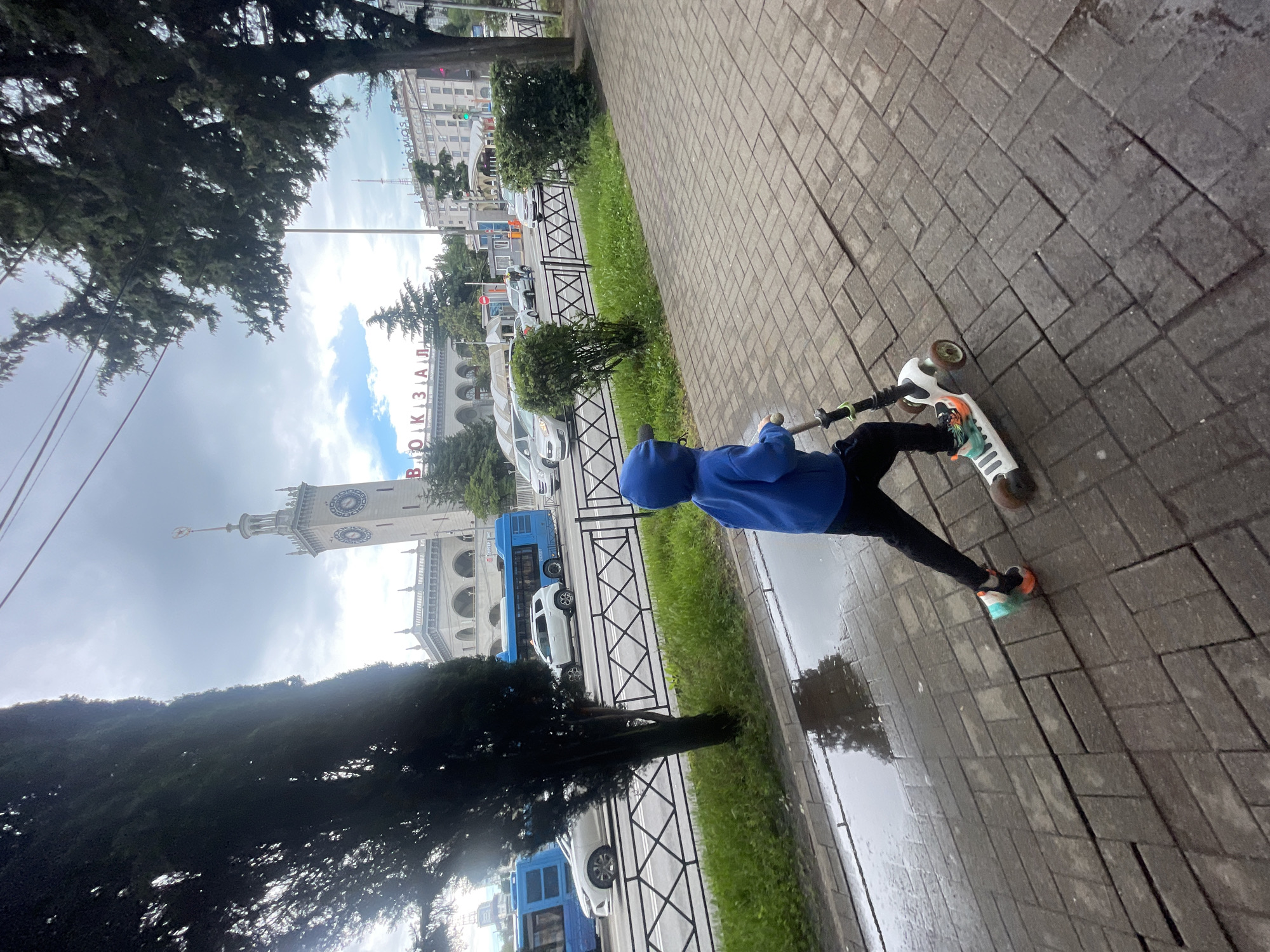 A full shot captures a child riding a scooter on a sidewalk. The child wears a blue hoodie and dark pants. The scooter is primarily white, with orange on the wheels and light green designs near the front.
In the background, across a street, is the Sochi railway station, with distinctive clock tower. Cars and buses are parked along the road. Trees frame the scene on the left, partially obscuring the view. The sidewalk is paved with rectangular grey blocks. Puddles of water are on the sidewalk. The sky is overcast, with visible clouds. The image is taken in Sochi, Russia.
