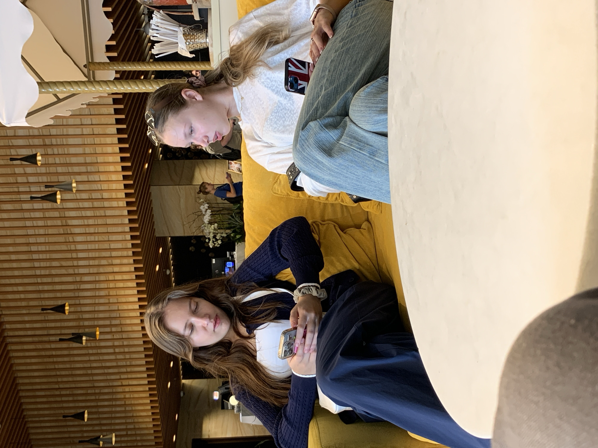 Two young women are seated on a yellow sofa in what appears to be a cafe or lounge area. The woman on the left is looking down at her mobile phone, which she holds in her hands. She is wearing a dark blue cardigan over a white top and dark blue pants. Her hair is long and brown. The woman on the right is looking to her left and speaking. She is wearing a white t-shirt and light blue jeans. Her hair is blonde and pulled back in a ponytail. Her phone, with a British flag case, is resting on her leg. In the background, a decorative wooden ceiling with pendant lights is visible, along with other patrons and cafe furnishings. The environment suggests an indoor public space in a city. There is no specific indication of the city being Moscow, Russia.