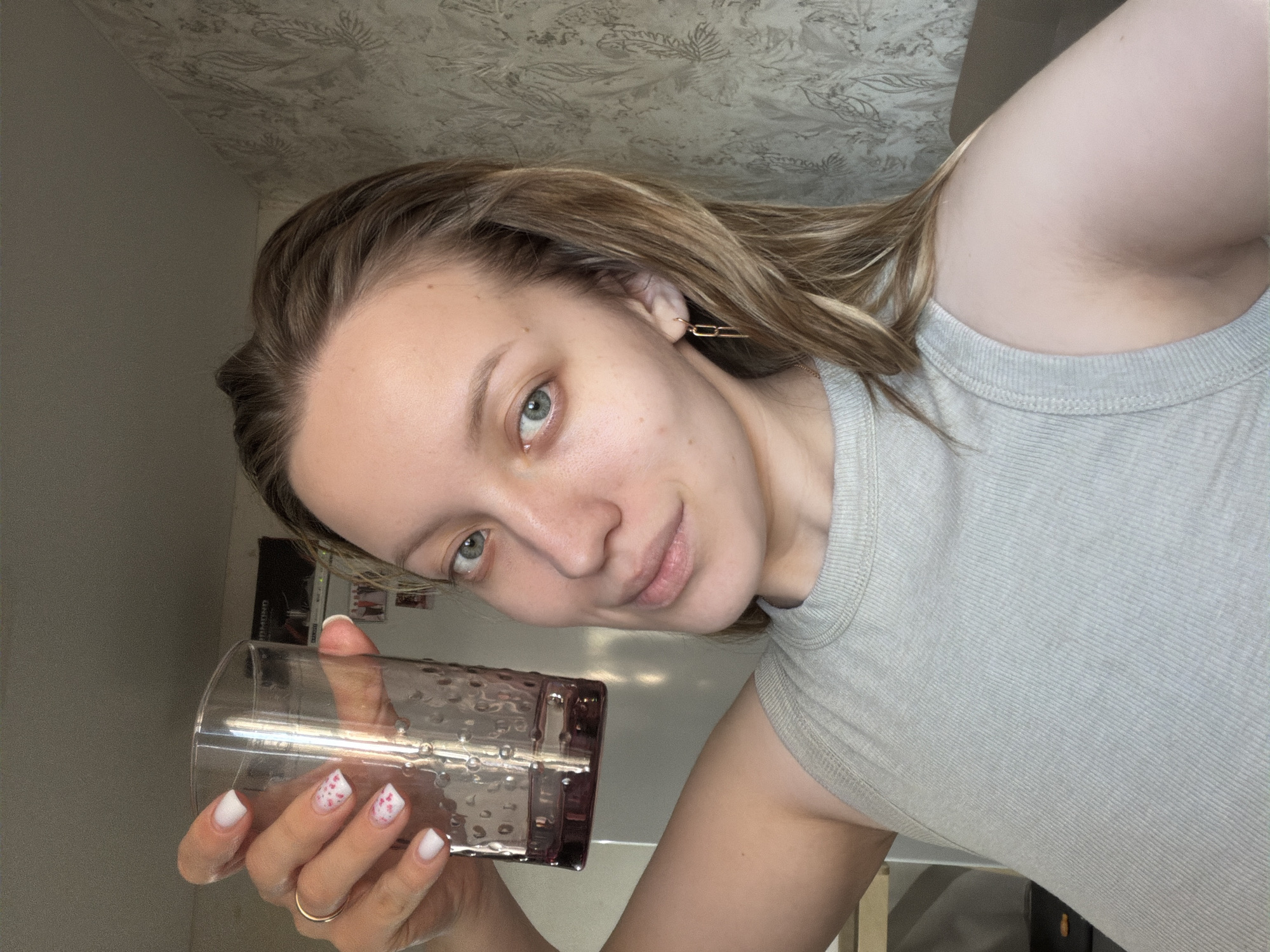 An adult female is depicted indoors in Reshetnikovo, Russia, holding a glass. The woman has light skin, blue eyes, and light brown hair. She is wearing a light grey ribbed tank top and a gold chain-link earring in her right ear. A gold ring is visible on her left ring finger. Her left hand, with white manicured nails featuring pink patterns on the ring and pinky fingers, holds a clear glass with a textured surface and a purple tint at the bottom, containing a small amount of liquid. She is looking directly at the camera with a slight smile. The background reveals a light-colored wall with patterned wallpaper, a white appliance (possibly a refrigerator) with papers or photos attached, and a dark rectangular object on top of the appliance. A light brown furniture edge is partially visible in the lower right background.