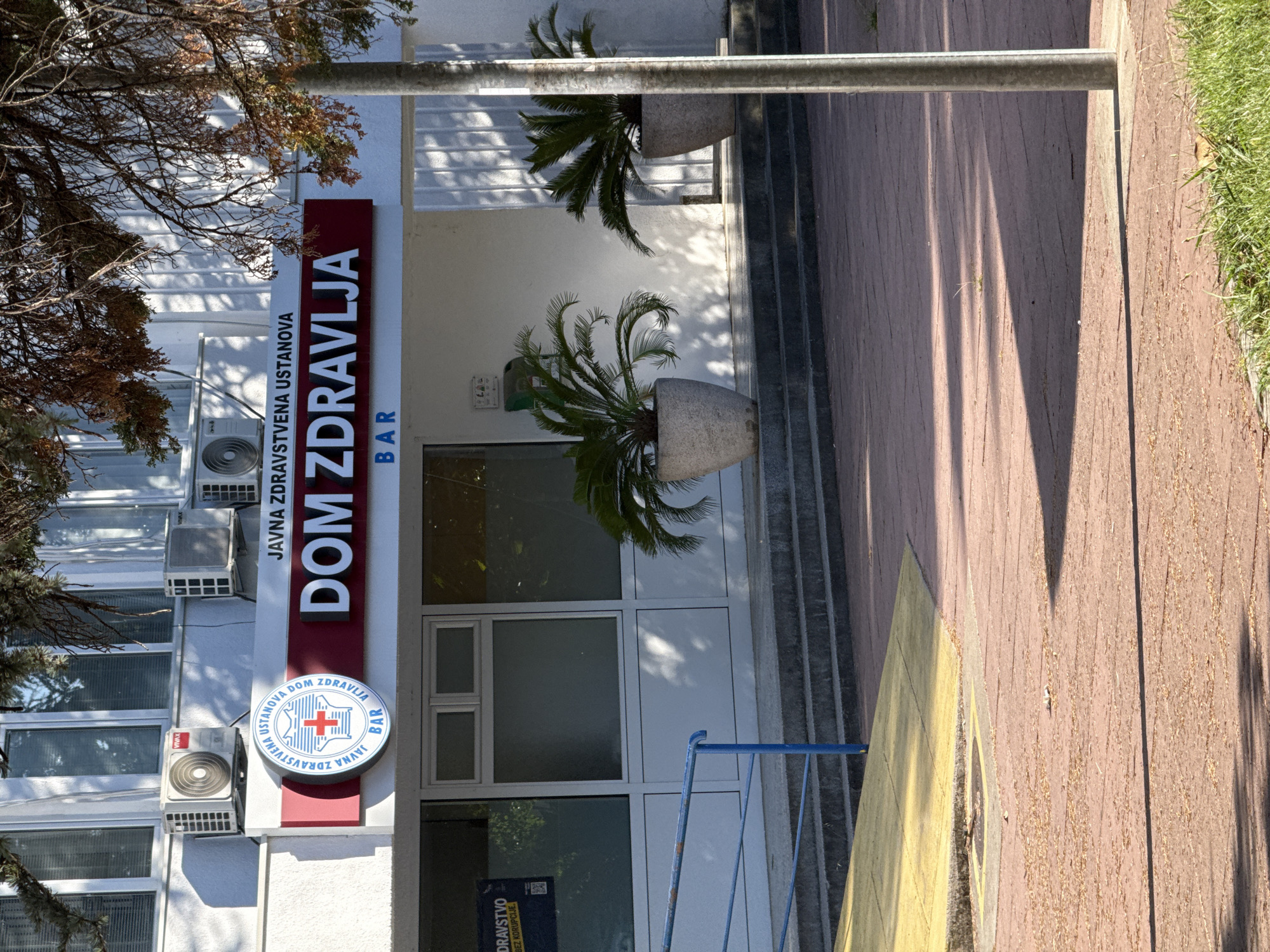 The image shows the exterior of a public health institution named "DOM ZDRAVLJA" in the city of Bar, Montenegro. The building is white with several windows and air conditioning units visible on the facade. A red sign above the entrance displays the name of the institution in large white letters, with smaller text above it and a circular logo to the left. The logo is blue and white with a red cross. Below the main sign, the word "BAR" is visible. In front of the building, there are two potted plants and a set of steps leading to the entrance. A yellow ramp is also visible on the left side, and a metal pole stands in the foreground on the right. The ground in front of the building is paved with reddish-brown tiles, and some dry leaves are scattered on the ground. The overall scene appears to be a daytime shot with sunlight casting shadows.