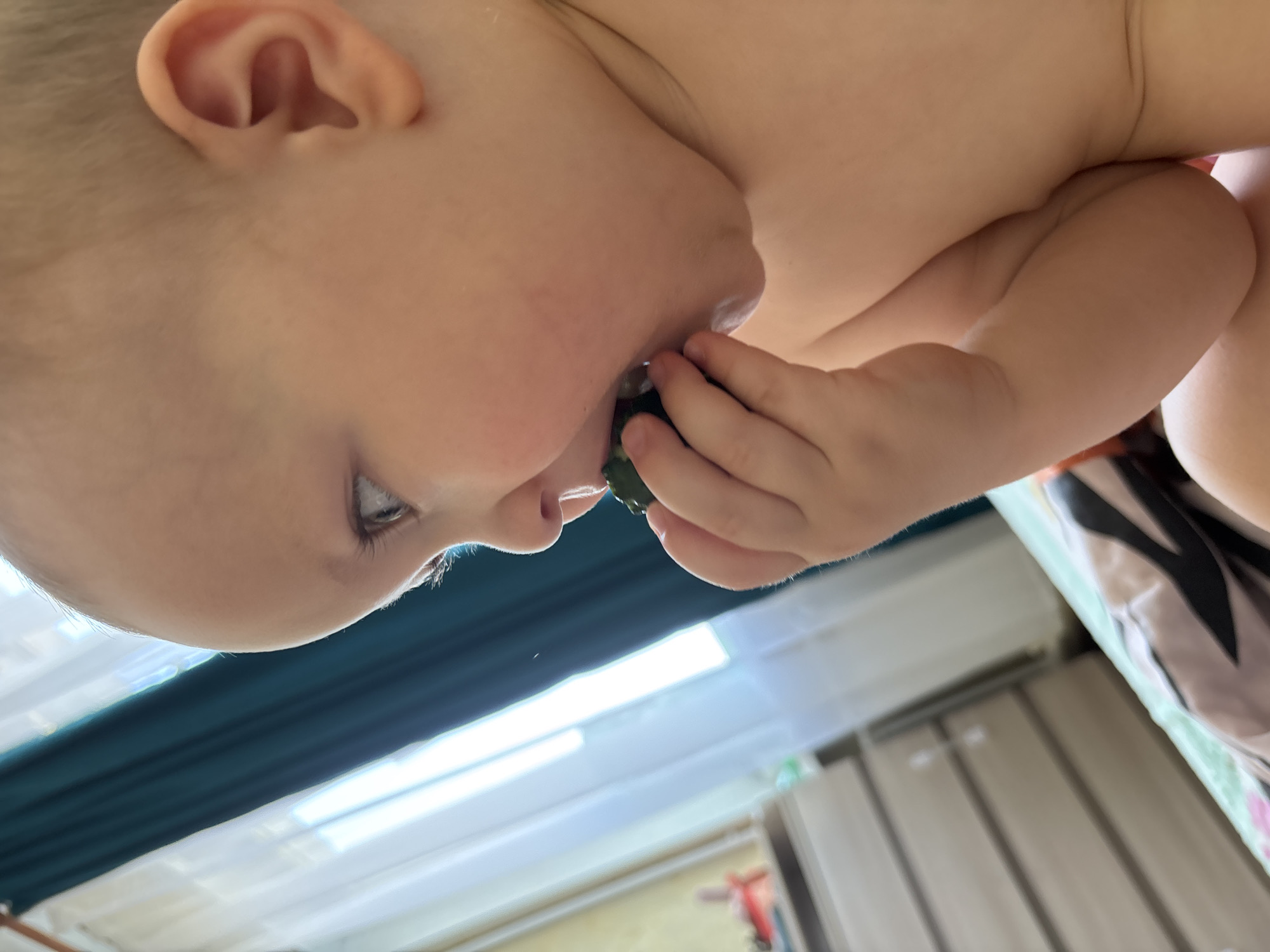 A close-up, profile view of a baby's head and upper body. The baby has fair skin and blonde hair. Their right eye is wide open, looking to the left. Their mouth is open, with a small, dark green object, possibly food, being held near their lips by their right hand. The baby is positioned in the foreground, against a backdrop that includes a teal curtain and a window with bright light. In the blurred background, a dresser with multiple drawers is visible, as is bedding. The image appears to be taken indoors. The location of this scene is not specified in the provided information, and therefore cannot be identified as being in Naberezhnye Chelny, Russia.
