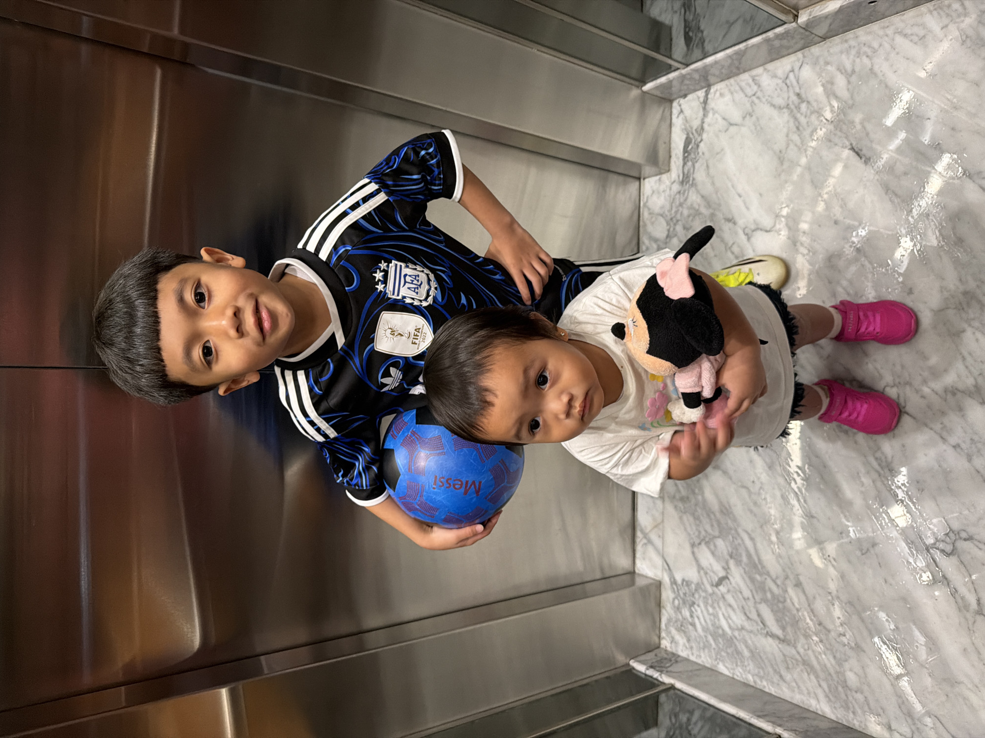 Two children, a boy and a younger girl, stand in an elevator with brushed metal walls and a marble floor. The boy, wearing a black and blue soccer jersey, holds a blue soccer ball with red markings. The girl, in a white t-shirt, clutches a stuffed toy. The scene appears to be an indoor location in Central Jakarta, Indonesia, likely within a residential or commercial building.
