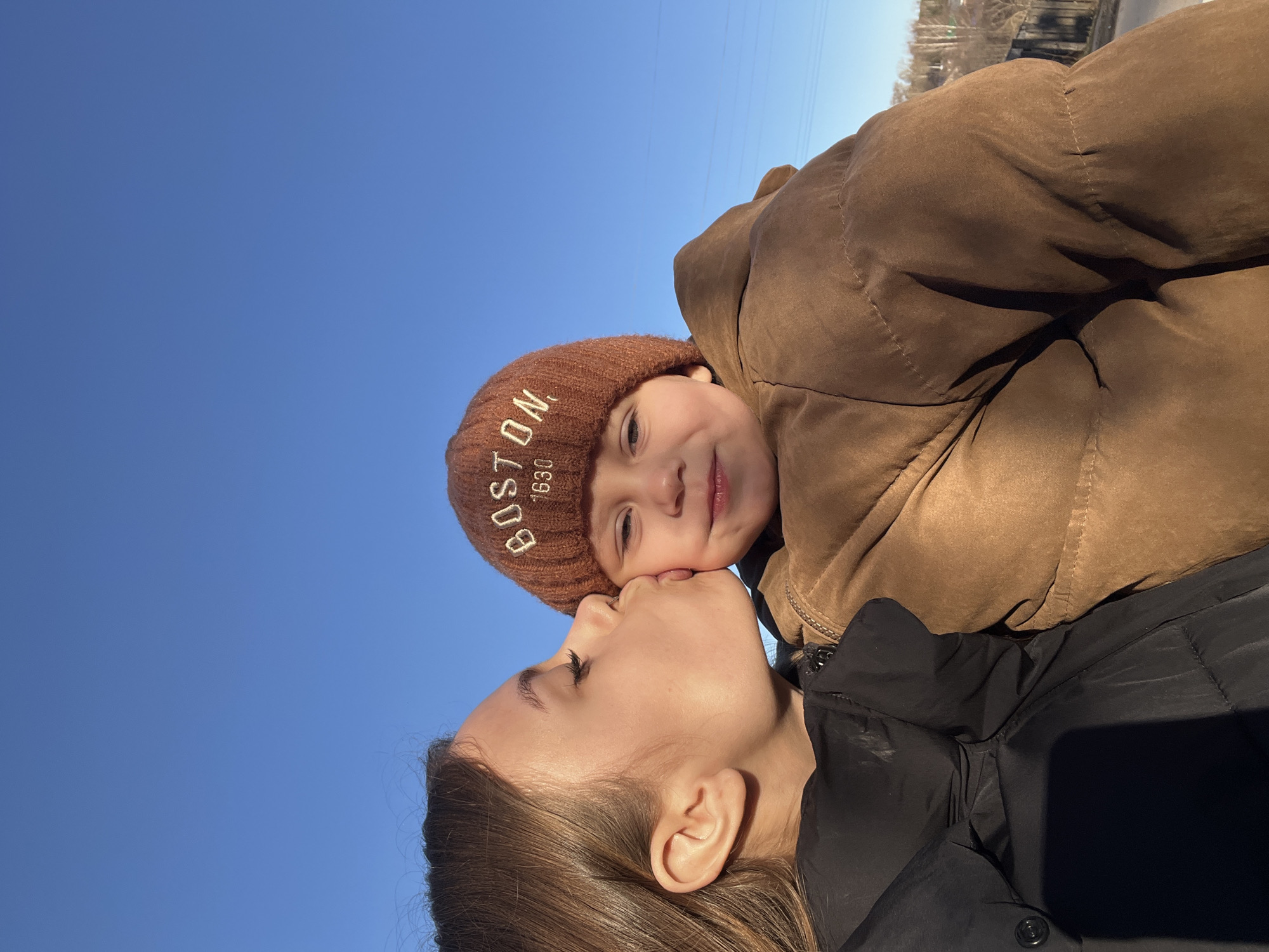 A photograph shows a woman kissing a young child against a bright blue sky. The woman has brown hair and is wearing a black jacket. The child is wearing a brown jacket and a brown knit hat with "BOSTON 1630" embroidered on it. The location cannot be determined from the image.