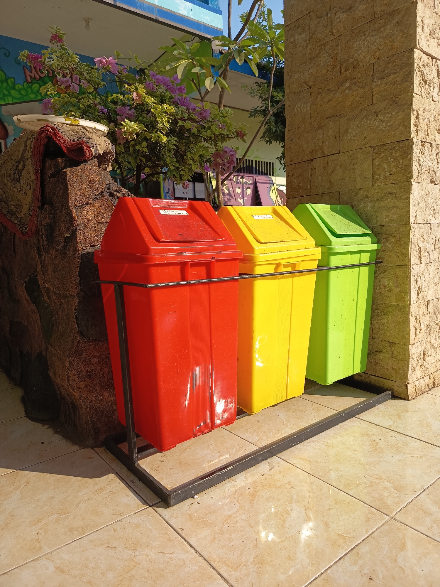 In Gebangan, Indonesia, three brightly colored plastic trash bins—red, yellow, and green—are lined up on a tiled outdoor surface. A large, textured, dark brown structure, possibly a decorative element or a natural rock formation, stands to the left of the bins. Behind the bins, a flowering plant with purple and pink blooms adds a touch of color to the scene. The background shows a building with a blue facade and a wall constructed of light brown stone blocks. The overall environment suggests an outdoor area, potentially a school or public space, bathed in natural sunlight.