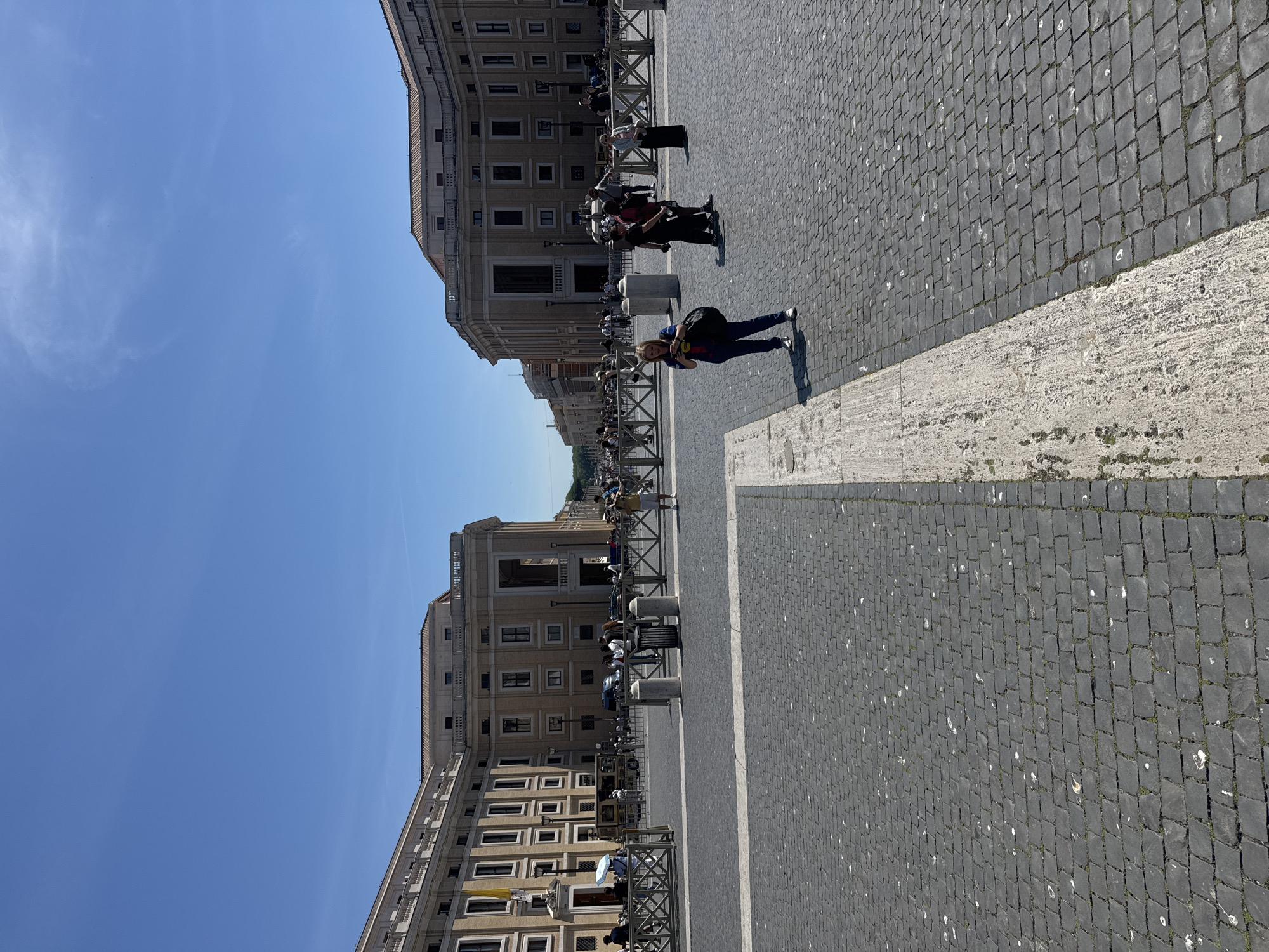The image captures a bright day in St. Peter's Square, Vatican City, Rome, Italy. The sky is a clear blue. The square is paved with dark cobblestones, marked by a wide, light-colored border. Buildings with classical architecture line the square. Numerous people are present, some standing behind metal barriers. A woman in blue jeans and a dark shirt stands in the foreground.
