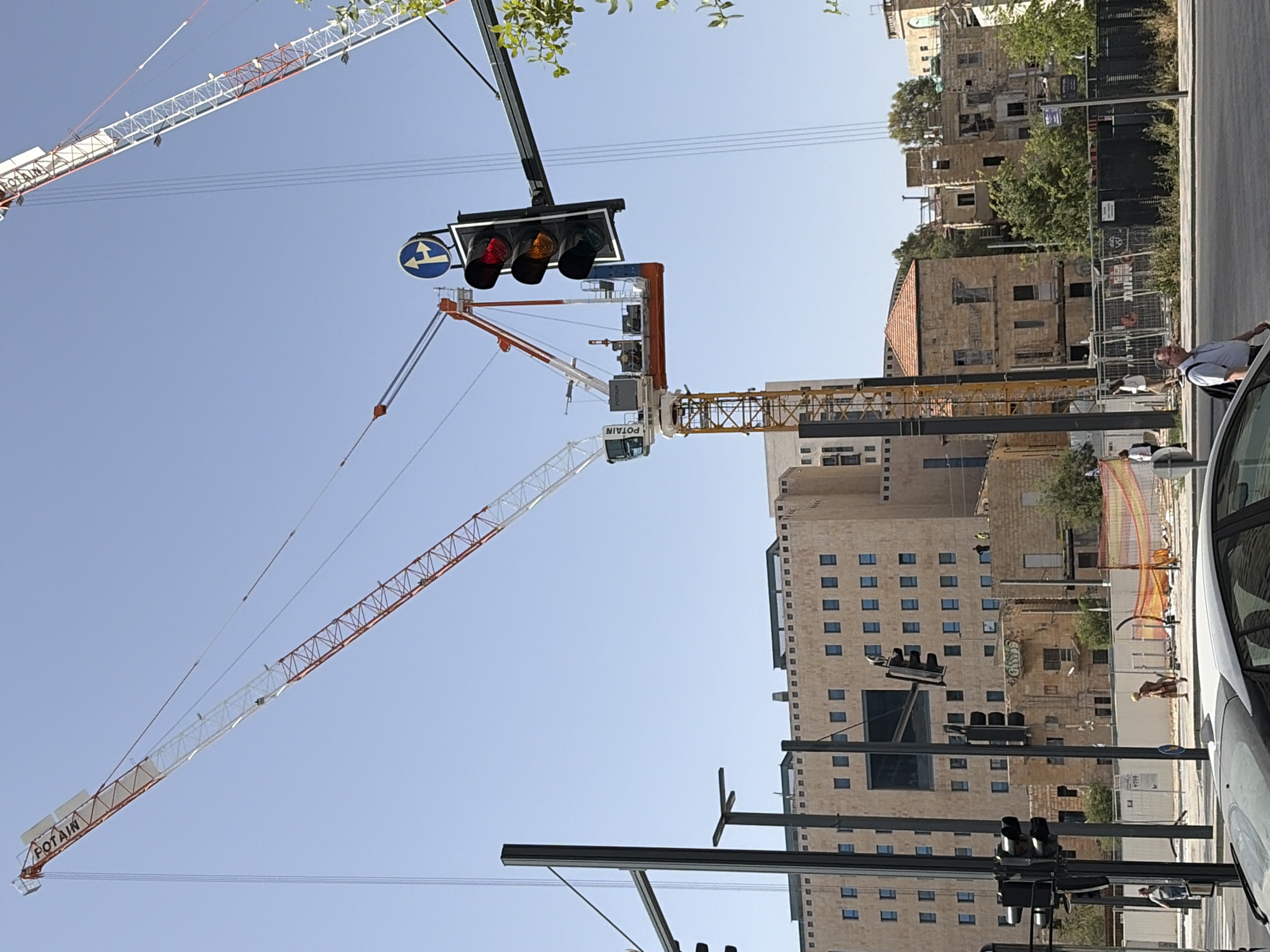 The image depicts a street scene in an urban area, likely a city, with prominent construction activities. The focus is on two large construction cranes dominating the skyline against a clear, blue sky. One crane is primarily white with some red accents, and the other is yellow and white. A set of traffic lights is visible, with red and yellow lights illuminated. Adjacent to the road, there's a modern building with numerous windows, constructed of light-colored stone. A smaller, older building with a red roof is also present. People are seen walking along the sidewalk, and a portion of a silver car is visible on the right edge of the image. The image suggests an area undergoing development or redevelopment.