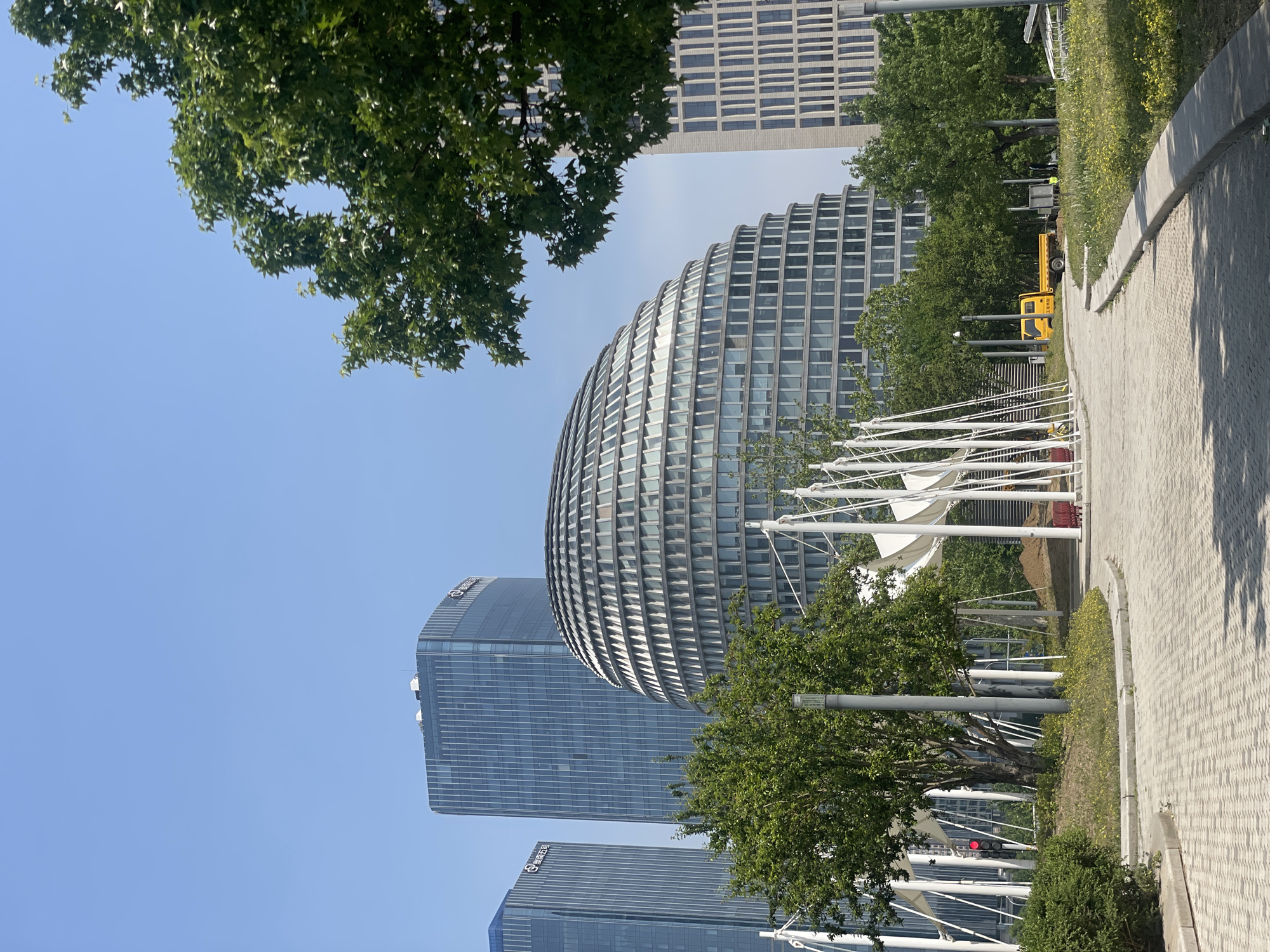 The image captures an urban scene in the Chaoyang District of China, featuring a mix of modern architecture and green spaces. A prominent, dome-shaped building with a grid-like facade stands among taller, rectangular skyscrapers. Trees partially obscure the view, adding natural elements to the cityscape. A paved walkway runs along the right side of the frame, suggesting a pedestrian area. In the background, there is a yellow truck. A traffic light is also visible. White structures, possibly part of a public art installation or architectural feature, are seen near the walkway. The sky is clear and blue, indicating a sunny day.