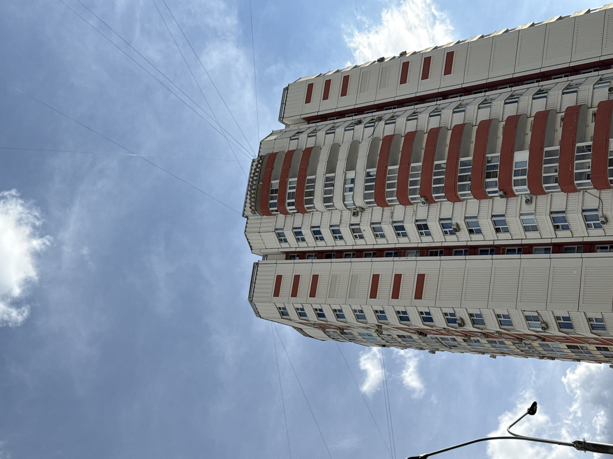 A low-angle, full shot captures a towering, multi-story apartment building against a bright blue sky with wispy white clouds. The building's facade is predominantly white with vertical ribbed detailing and prominent horizontal bands of red. Many windows are visible, some with visible air conditioning units below them. Curving balconies with red railings punctuate the building's shape. Several power lines crisscross the sky above the building. To the left, a black lamppost with a curved arm is partially visible. The image appears to have been taken in Lyubertsy, Russia.