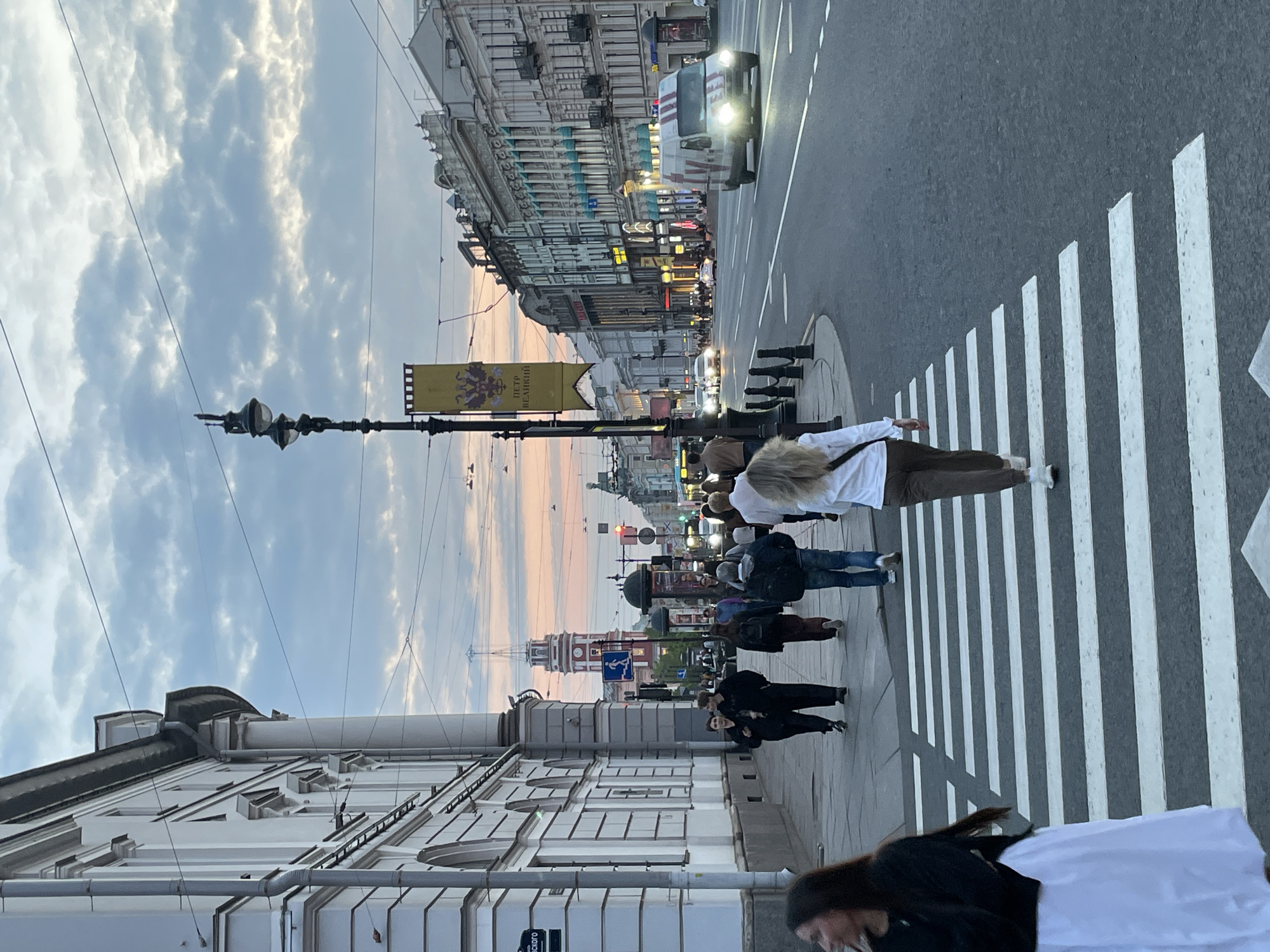 The image captures a street scene in Saint Petersburg, Russia, during daylight. Pedestrians are crossing a zebra-striped crosswalk. Buildings line the street, with a view towards a tower in the distance. A street lamp and a banner with text in Russian are visible. A vehicle is driving on the road. The sky is partly cloudy.