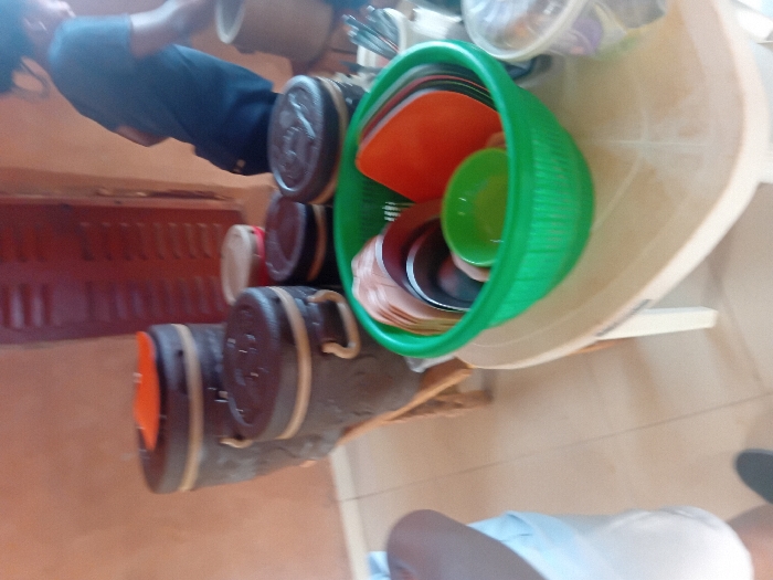 This image depicts an indoor scene within a commercial establishment, likely an eatery or shop, in Ejule, Nigeria. The foreground shows a light-colored tiled floor. On the left, the lower torso and legs of an individual wearing a light-colored top and dark footwear are partially visible. A light-colored, possibly beige or off-white, plastic table occupies the mid-ground, holding a green plastic colander filled with various dishes, including cream-colored plates, a dark plate, a small green bowl, and an orange rectangular item. To the right of the colander, a transparent plastic container with a lid is visible. Behind the table, several dark brown cylindrical insulated containers or food warmers with lighter bands and handles are arranged; one taller unit has an orange item on its lid. In the background, a person wearing a dark blue t-shirt is partially visible from the waist up, their head and upper body obscured as they appear to be leaning or reaching towards an object out of frame to the right. The background wall is a light brown or orange hue, featuring a prominent reddish-brown wooden louvered shutter or window.