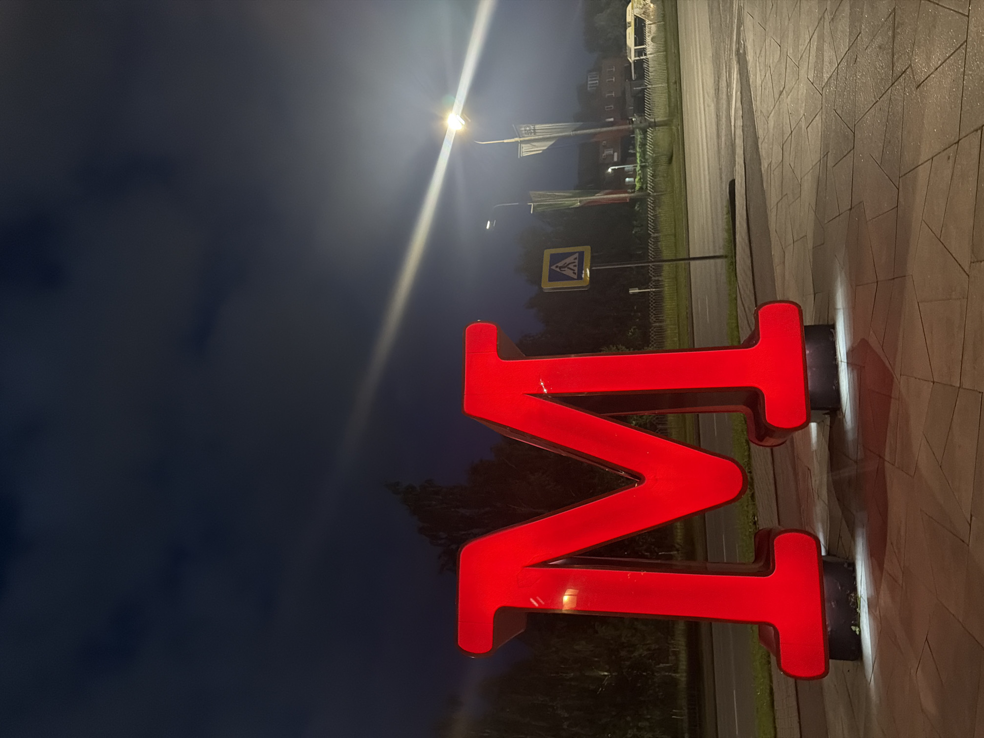 A large, red, illuminated letter "M" stands prominently on a paved area at night. The letter is a three-dimensional structure, likely a sign, with internal lighting that casts a glow. In the background, there is a road, a pedestrian crossing sign, lampposts with flags, trees, and a building with windows. A white bus or van is partially visible on the road in the distance. The sky is dark, indicating it is nighttime, with some clouds and light streaks from artificial lighting. The ground is paved with geometric stone tiles. The scene appears to be an outdoor public space, possibly a street or a plaza, in Naberezhnye Chelny, Russia.