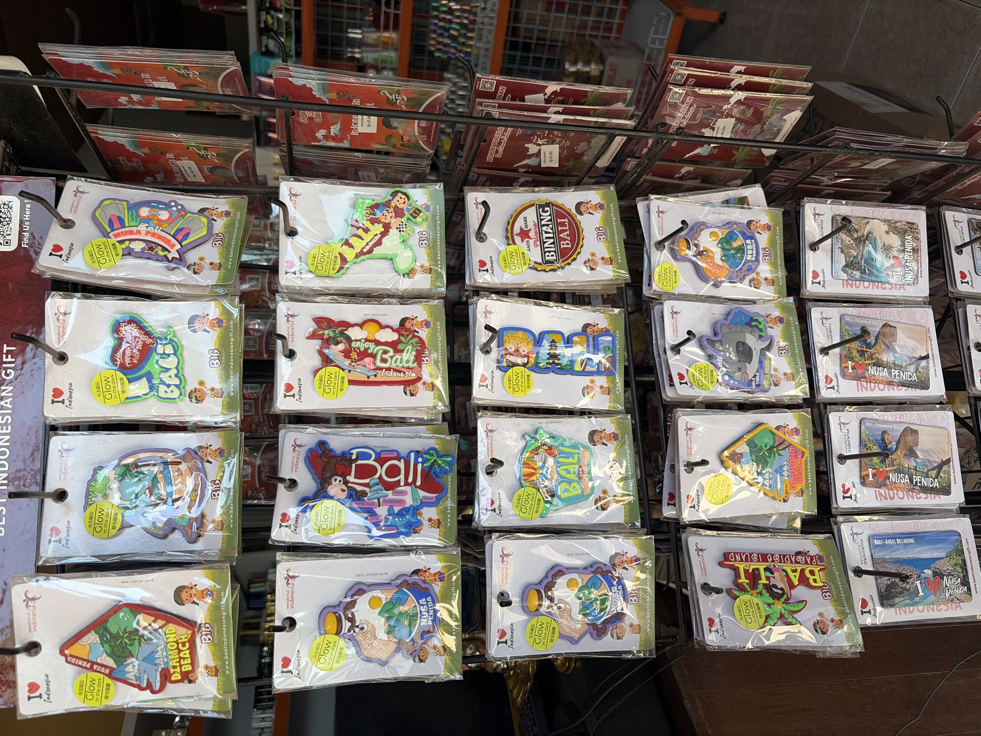 The image shows a retail display of souvenirs in Nusa Penida, Indonesia. The souvenirs are primarily magnets, most of which feature the "I Love Indonesia" logo. The magnets are packaged in clear plastic and hung on black metal display racks. Designs on the magnets include scenes of beaches, the text "Diamond Beach," "Nusa Penida," and "Bali." Some magnets have Japanese text that translates to "Glow in the dark." Several of the magnets are shaped like dinosaurs or cartoon characters. The display also includes some flat souvenir cards with similar designs. In the background, shelves stocked with other products, likely more souvenirs or general store items, are visible, along with a partial view of an orange shopping cart. The lighting suggests an indoor setting, possibly a gift shop.