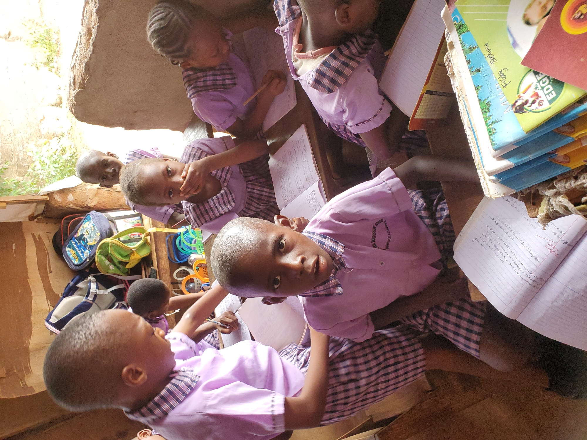 The image captures a group of young children engaged in a learning activity within what appears to be a simple classroom in Ukwu-Abwa, Nigeria. Several children are visible, mostly wearing light purple shirts with plaid collars and cuffs, indicative of school uniforms. Their hair is closely cropped or styled in braids.
The children are seated at desks or benches, diligently working in notebooks with pencils. One child in the foreground looks directly at the camera with an open expression, while another nearby covers their mouth. Stacked books, some with colorful covers, are visible on a desk to the right, with one clearly showing "Friday School" and "EDGE" on its cover. School bags and what appear to be colorful learning toys or aids are scattered in the background, suggesting a dynamic learning environment.
The setting features rough, unadorned walls, possibly made of stone or concrete, and natural light filters in from an unseen opening, indicating it is daytime. Outside, some greenery is visible, adding to the rustic feel of the location. The overall atmosphere suggests focused learning despite the simple surroundings.
Visible text includes "WATESSOR SCHOOL" and "NOWELLE EZLINNAKA" on a child's uniform. In open notebooks, various handwritten lines are discernible, including phrases like "Risks are Ring...", "COWISE SOMEtting bad to happen...", "Something a risk factor," "pre existing," and "Look and serve Foo." There are also numbers like "260126" and words like "sefe" and "Friday Sochina" (likely "School").