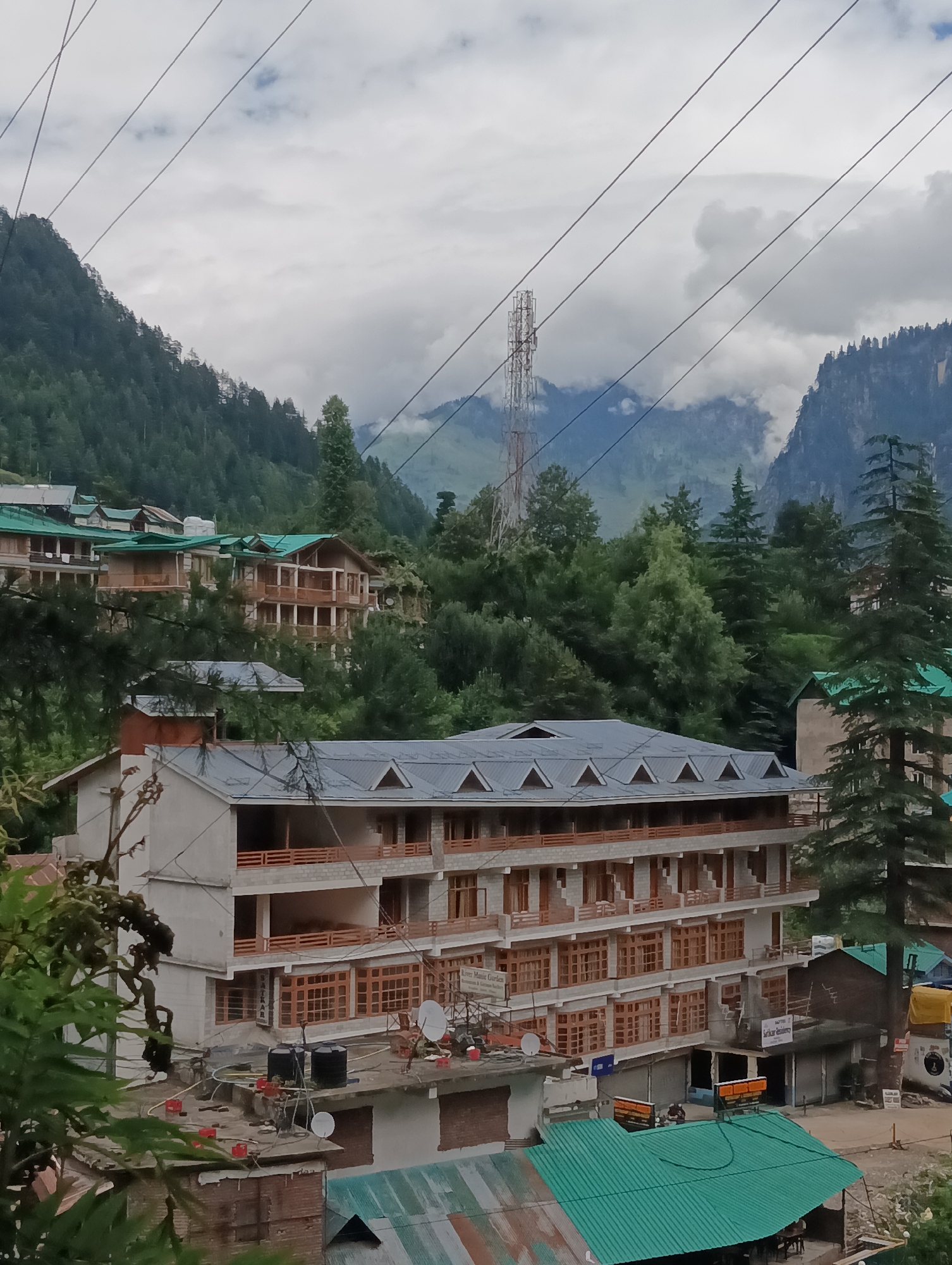 The image depicts a view of buildings and surrounding landscape in Manali, India. In the foreground, a multi-story building with a light-colored facade and numerous wooden-framed windows is visible. This building appears to be a hotel or guesthouse, with balconies and external staircases. Several other buildings with green-tiled roofs are also present, nestled among trees.
The background features lush green hillsides covered in dense forest. In the distance, a tall telecommunications tower stands against a cloudy sky, with mist or clouds obscuring the upper parts of the mountains. Electrical wires are strung across the sky, adding to the urban or semi-urban feel of the scene. The overall impression is of a mountainous town with a mix of natural scenery and human habitation.