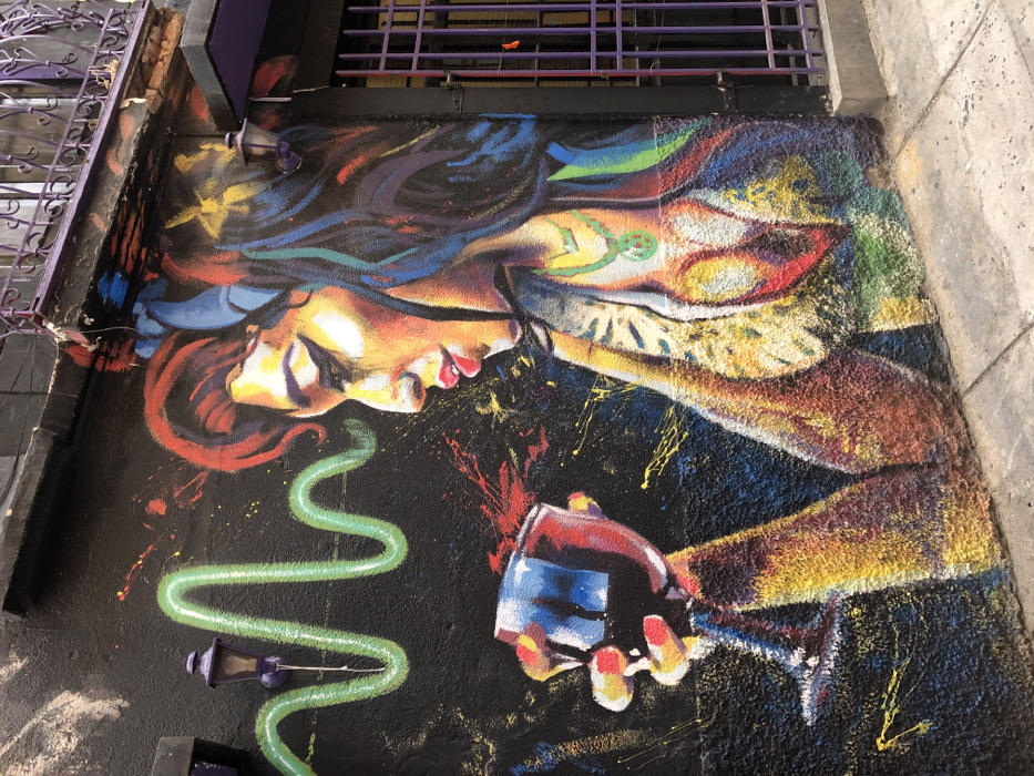 A vibrant, colorful mural features a woman lying horizontally, her face illuminated with warm tones, framed by swirling hair in shades of red, blue, and black. She seems to be holding a glass, painted in bright red, blue, and white, above a textured black background. A lime-green, serpentine shape meanders near the glass, punctuated by small, purple fixtures. The painting is positioned on a rough surface, flanked by a light-grey wall and a dark-grey iron structure.