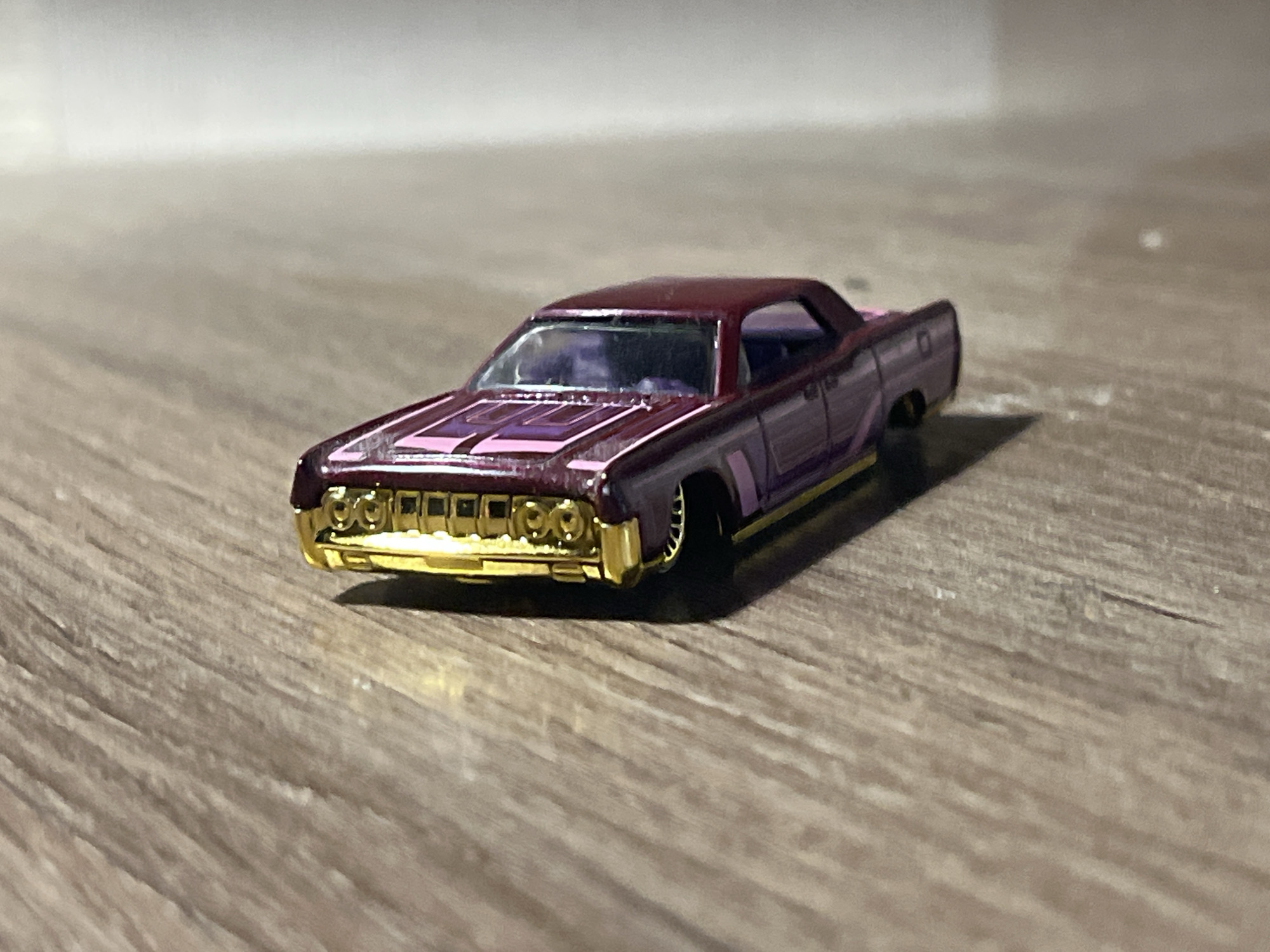 A close-up, low-angle shot captures a maroon die-cast toy car with pink racing stripes on its hood and a gold grille, resting on a light brown wooden surface. The car is positioned at an angle, with its front facing left, showcasing its detailed gold bumper and headlights. The background is a blur of light grey and white, suggesting an out-of-focus environment. The image does not provide any information about the city of Svietlahorsk, Belarus, or any inhabitants.