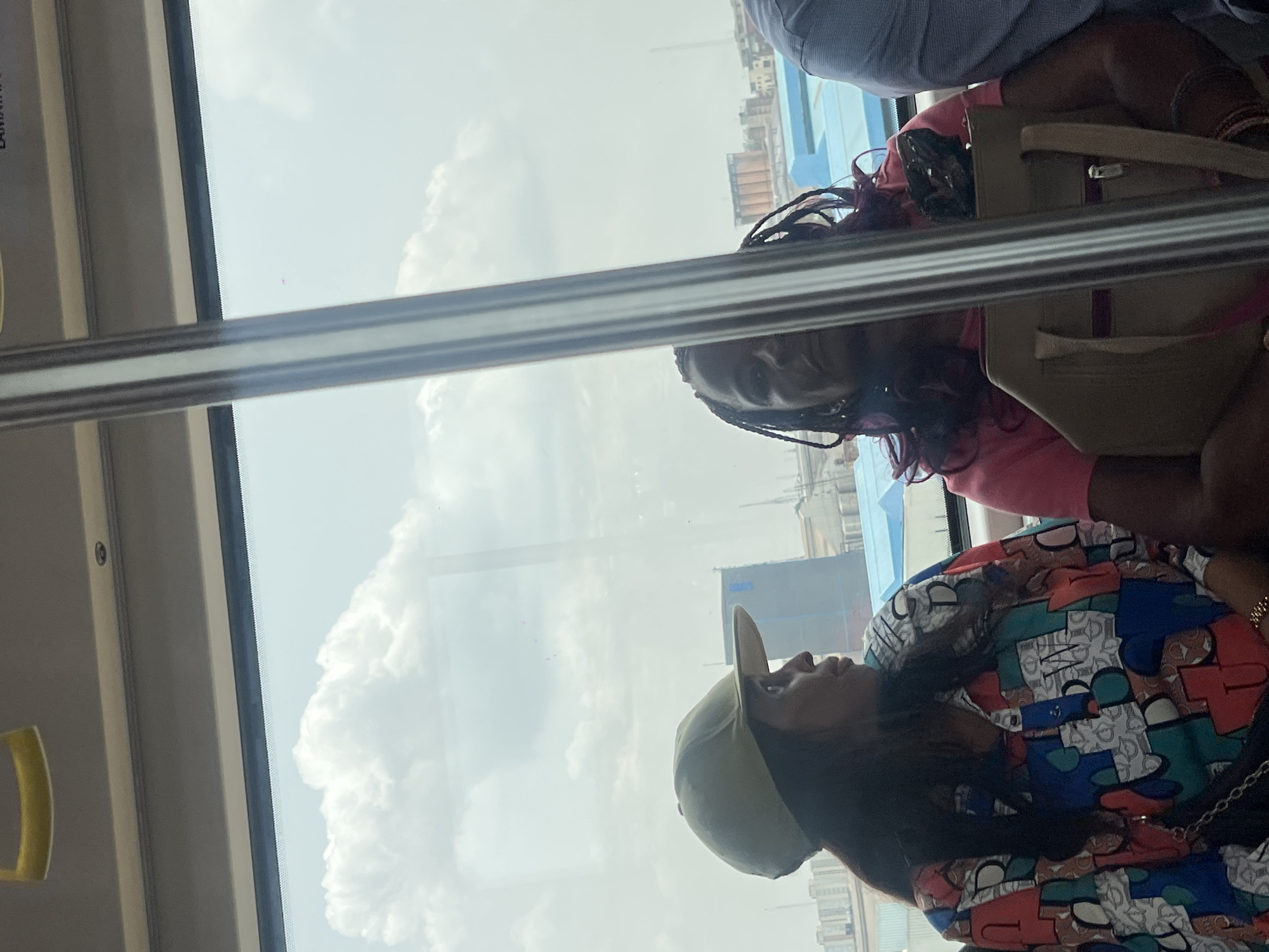 Two women are seated on a public transport vehicle. One woman wears a pink shirt, braids with reddish tints, and has a beige handbag. The second woman wears a colorful patterned shirt and a khaki baseball cap. The window behind them offers a view of Aqani, Nigeria; city buildings are visible in the background along with clouds. The interior of the vehicle is visible in the foreground.