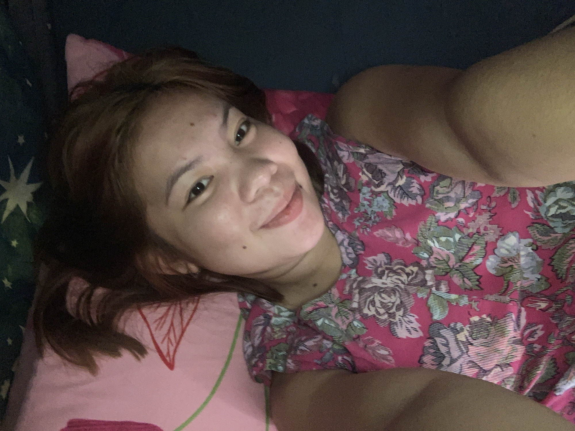A young woman with brown hair and fair skin is lying down on a pink pillow with a floral pattern. She is wearing a pink top with a floral pattern as well. She is smiling at the camera and her eyes are open. The background is dark, possibly a bedroom. The image appears to be a selfie taken by the woman. The image is not located in the city of Kiblawan, Philippines.