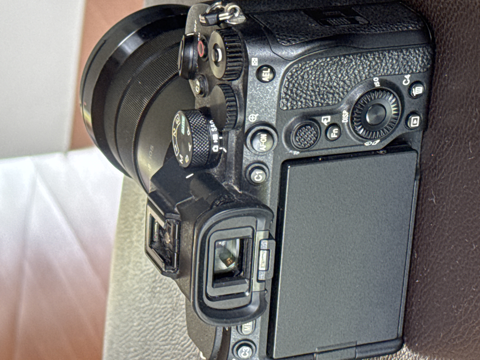 The image shows the back and top of a black Sony mirrorless camera with a lens attached. The camera is resting on a textured surface that appears to be a brown leather-like material. The camera has a viewfinder, a flip-out LCD screen, various buttons and dials, including a mode dial with settings like "M," "A," "S," and "AUTO," and a multi-selector dial with labeled functions such as "AF-ON," "AEL," "Fn," "DISP," and "ISO." There is also a hot shoe mount at the top. The background consists of a textured surface, possibly a table, with soft lighting. The image does not contain any people or depict any specific location in Ko Samui District, Thailand. It is a close-up product shot of a camera.