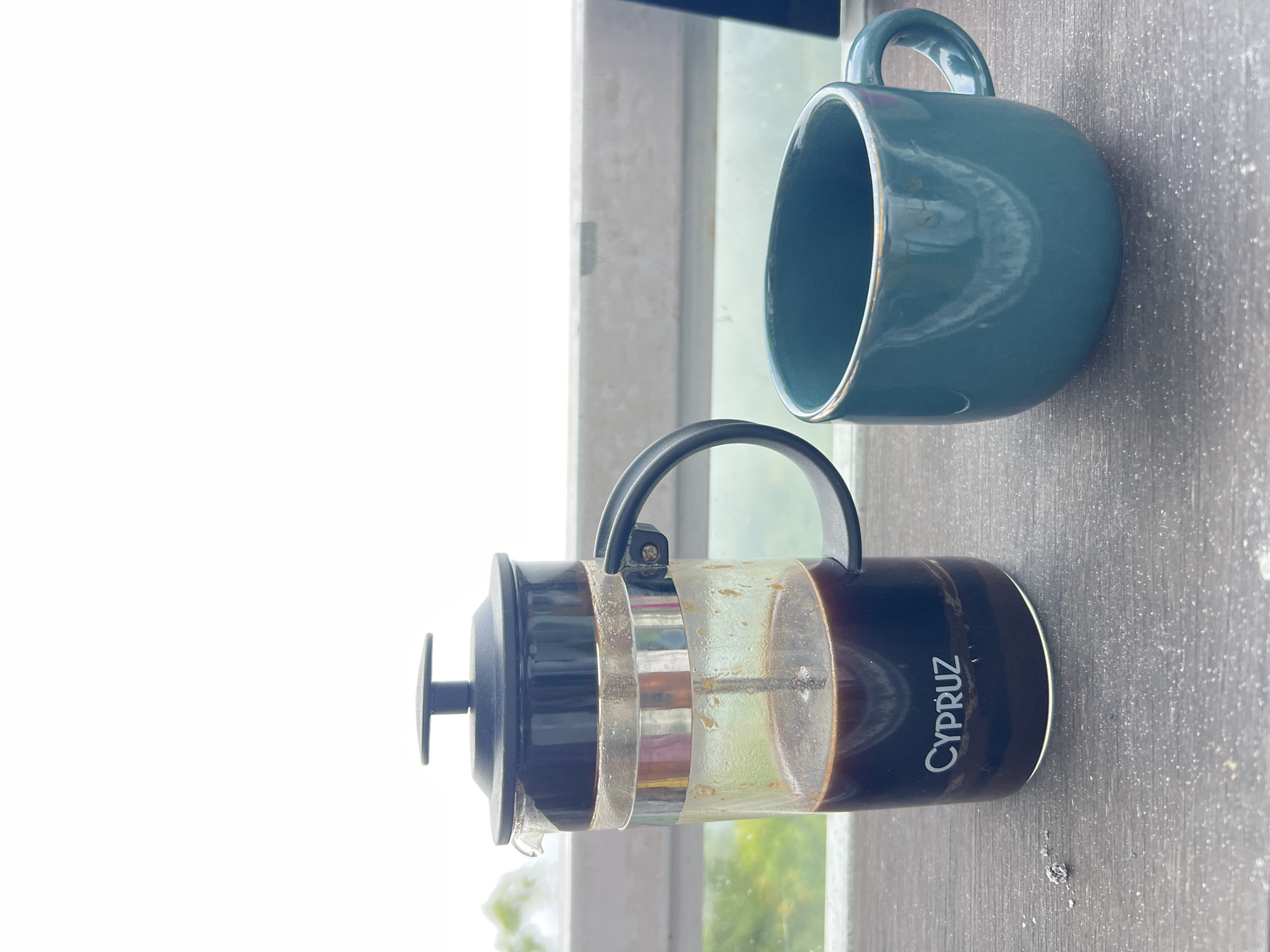 A French press filled with dark coffee sits on a wooden table next to a teal mug. The French press has a black lid and handle, and the glass container shows coffee grounds at the bottom. The word "CYPRUZ" is visible on the glass. The mug is rounded with a gold rim. In the background, a blurred green landscape is visible through a white railing, suggesting an outdoor setting, possibly a balcony or terrace with a view, consistent with the location in Kintamani, Indonesia, known for its scenic highlands and coffee plantations. The lighting is bright, suggesting it is daytime.