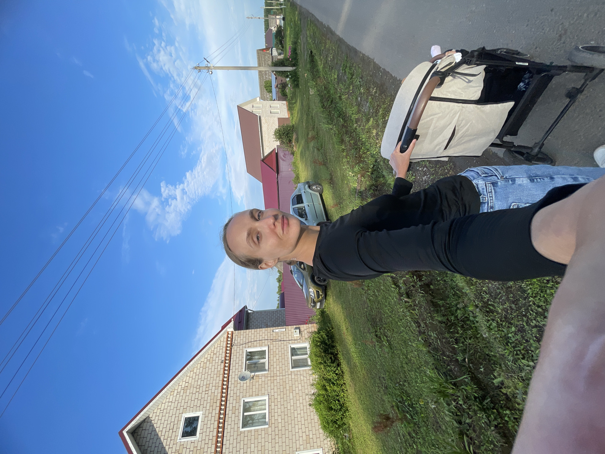 A woman, facing the camera with her right side angled away, walks down a paved road in a rural setting in Podshivalovo, Russia. She is pushing a beige and black stroller with her right hand. She wears a black long-sleeved shirt and blue jeans. Behind her, to the left, is a two-story brick house with a red roof. A green car is parked in front of a purple fence to the right of the house. Further down the road, more houses and a utility pole are visible under a blue sky with scattered clouds.