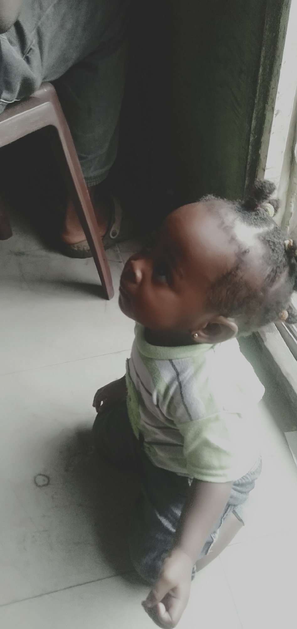 A young Black child is kneeling on a tiled floor, looking upwards and to the right. The child has dark skin, black braided hair with yellow bands, and is wearing a light green and white striped t-shirt and dark shorts. The child has a small earring in their right ear. The child is positioned in the foreground of the image.
Behind the child, a dark wooden chair is visible. To the left of the chair, the legs of an adult wearing dark trousers and light-colored sandals are partially visible, suggesting they are seated. The background is dark and indistinct, with a window frame visible on the right side of the image, suggesting the scene is indoors near an opening. The floor appears to be white or light-colored tile. The lighting suggests it might be daytime, with light coming from the window. The image was taken in Shangisha, Nigeria.