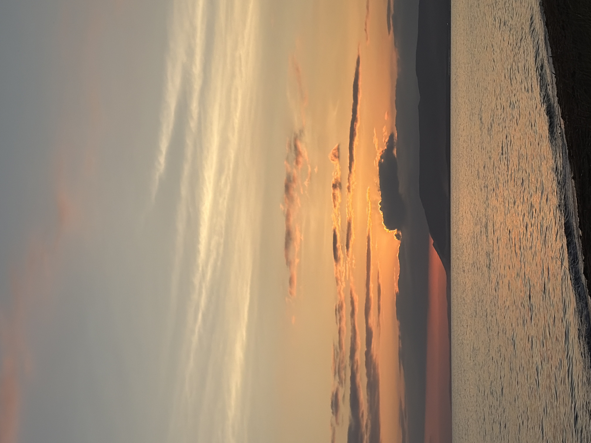 The image depicts a serene sunset over a body of water, with a mountainous silhouette on the horizon. The sky displays a gradient of colors, from a pale blue at the top to warm oranges and pinks closer to the horizon, interspersed with wispy clouds. The water reflects the sky's colors, creating a shimmering surface. A dark, silhouetted shoreline with a few blades of grass is visible in the foreground. The scene suggests a natural landscape, possibly a lake or sea. This location is identified as Ozerskoe, Russia. There are no people or discernible objects in the water or on the shore.