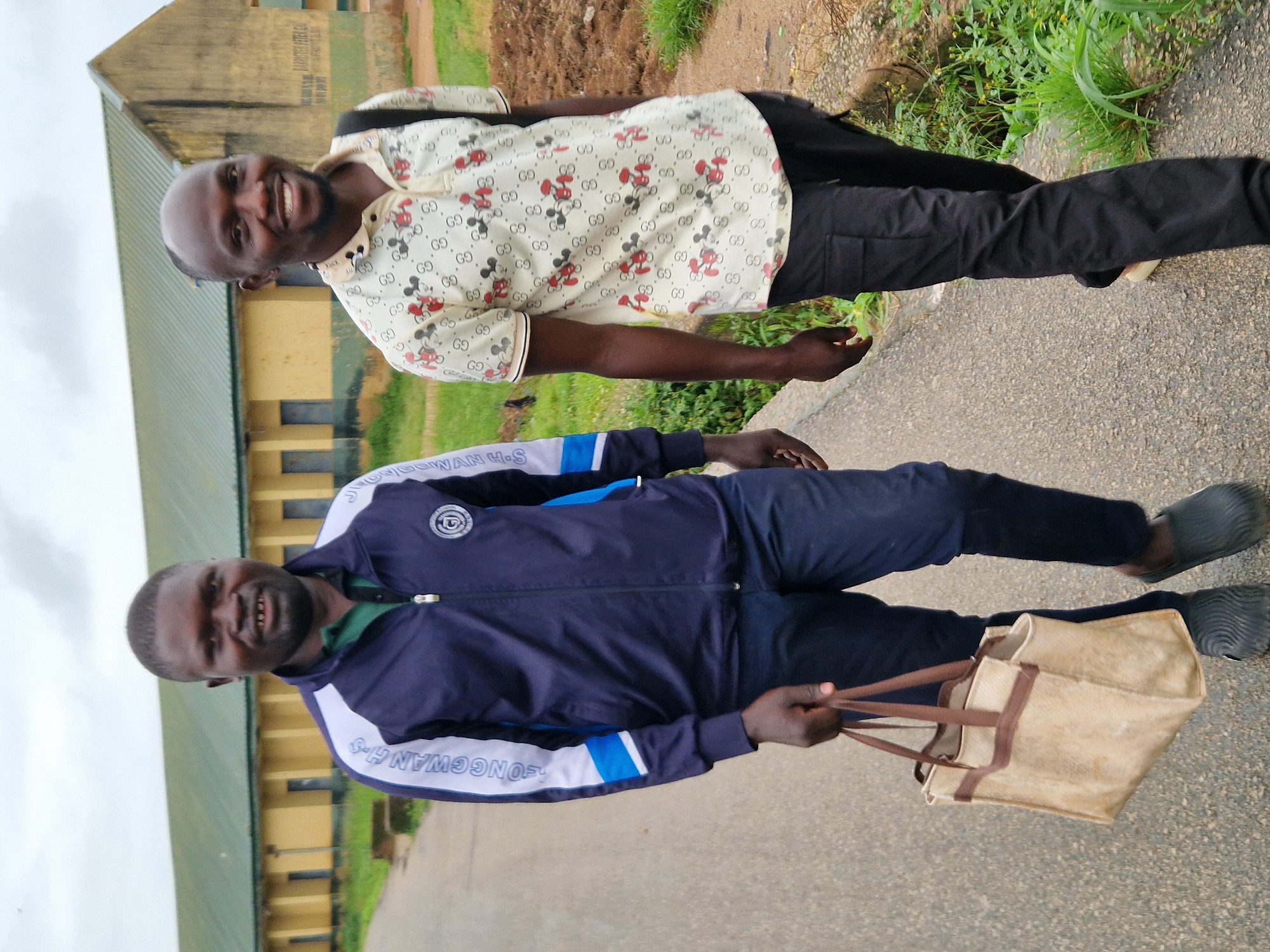Two adult males walk towards the camera on a paved road. The male on the left wears a dark blue jacket with white and blue accents on the sleeves, a green collared shirt, and dark blue pants. He is carrying a light brown canvas bag in his left hand. The male on the right wears a light-colored short-sleeved shirt with a pattern of Mickey Mouse heads and interlocking "G" symbols, and black cargo pants. They are walking in front of a long, yellow building with a green roof. The building appears to be a school or institutional building. Green vegetation and some disturbed earth are visible to the right of the road. The sky is overcast. The location is identified as Kabong, Nigeria.