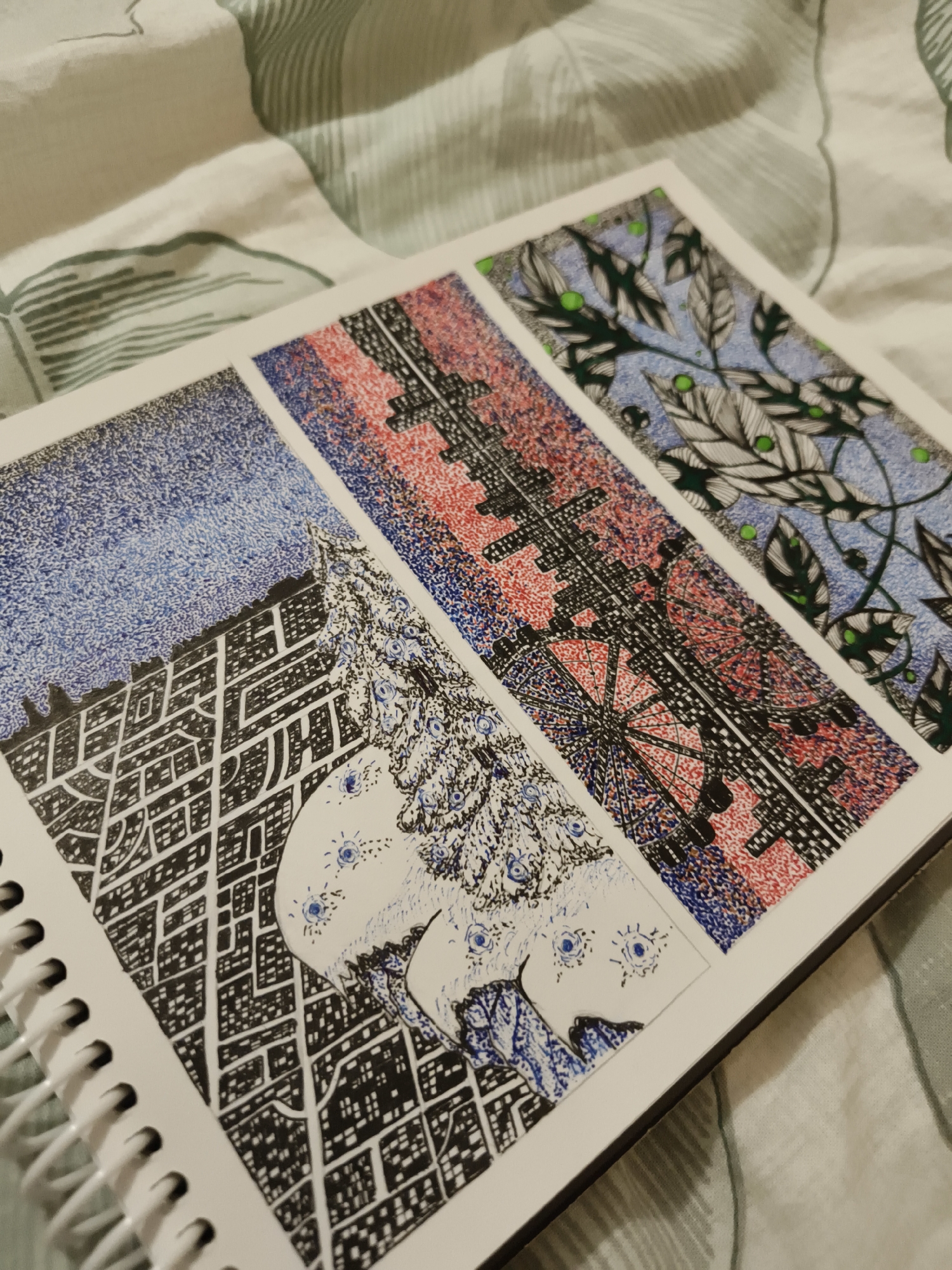 The image shows three framed drawings on a white paper in a notebook. The drawings are in a vertical format.
The leftmost drawing depicts a cityscape with a starry sky above. The city is rendered in black and white with a detailed grid-like pattern representing buildings and streets. A white, organic shape with blue accents, possibly representing a figure or an abstract form, is superimposed on the cityscape.
The middle drawing features a dark, intricate structure with red and blue stippling. This structure resembles a Ferris wheel or a similar mechanical object. The background is filled with a mix of red and blue dots.
The rightmost drawing shows a pattern of leaves and berries against a light blue background. The leaves are depicted in black and white with green dots representing berries.
The notebook is lying on a fabric surface with a green and white pattern. There is no indication of the city of Kurgan, Russia, in the image.