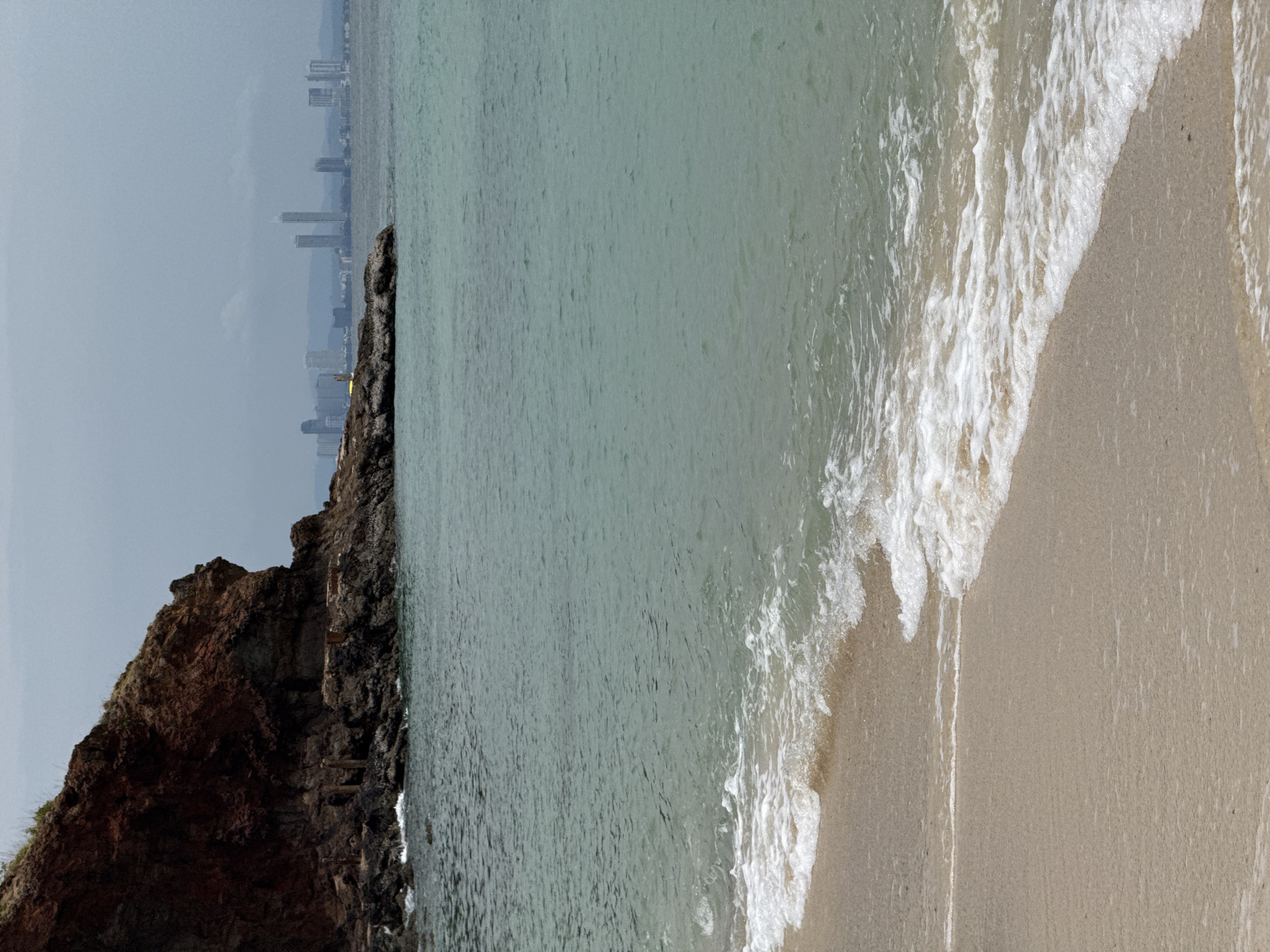 A scenic view of a beach with gentle waves is depicted. A rocky outcrop occupies the left side of the image, standing in contrast to the water. In the distance, a skyline of a city is visible, implying an urban area close to the coast. The location is Pattaya City, Thailand.