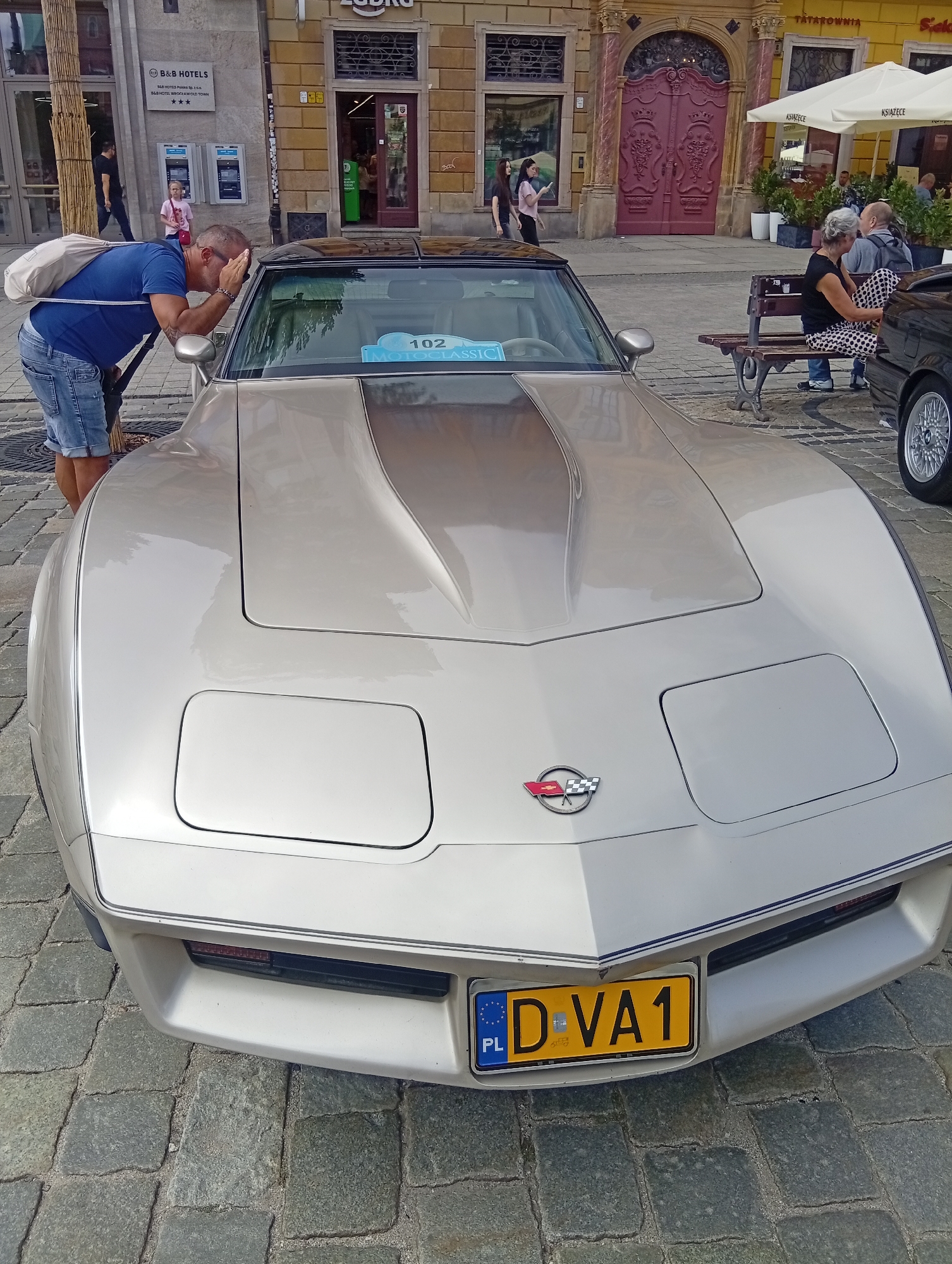 The image depicts a man in a blue t-shirt and denim shorts bending over the hood of a silver Chevrolet Corvette. The man has a light-colored bag slung over his shoulder. The car has a license plate reading "D-VA1" with the European Union emblem and the country code "PL" for Poland. In the background, there are buildings with storefronts, a woman with gray hair and a man sitting on a bench, and a young girl. The setting appears to be a public square with cobblestone paving. The architectural style of the buildings, particularly the ornate door with elaborate carvings and the signage like "B&B HOTELS," suggests a location in Wrocław, Poland, possibly in the Old Town area. The overall scene captures a moment at what appears to be a car exhibition or event in a European city.