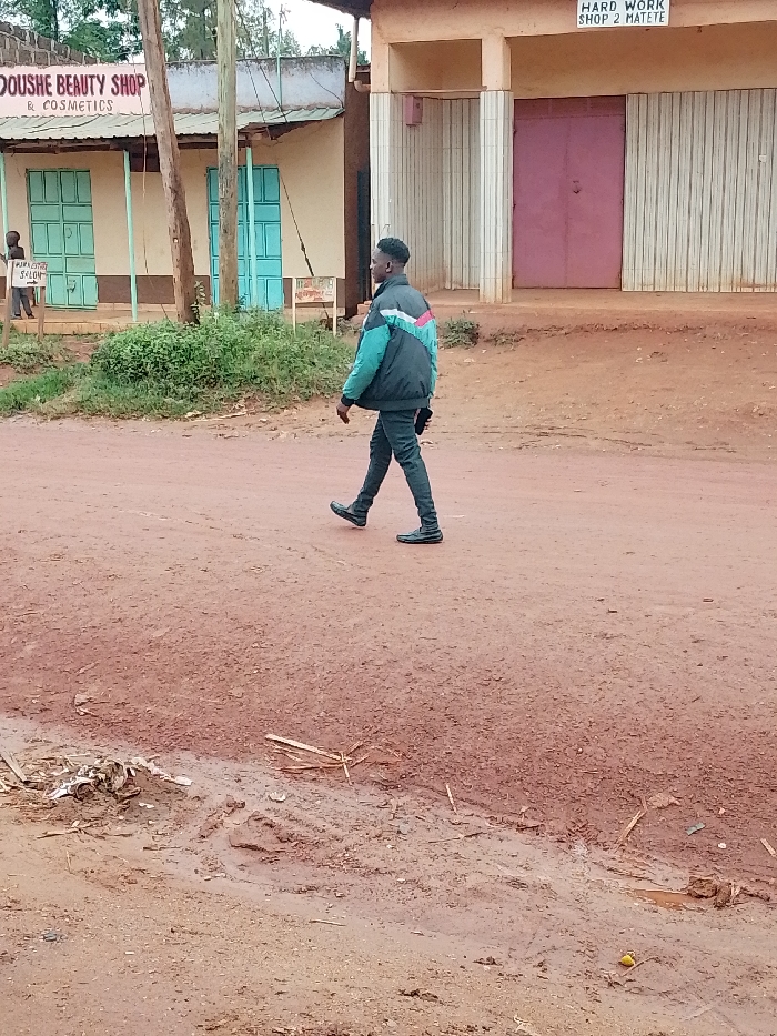 The image shows a man walking on a dirt road in front of several buildings, all of which are painted in shades of beige and brown. There are two buildings on the left side of the road, with signs indicating a beauty shop and a salon. The sign on the building on the right says “Hard Work Shop 2 Matete”. The man is walking on a dirt road that appears to have been recently rained on. He is wearing black jeans, black shoes, and a black and turquoise jacket. There are trees and bushes in the background. This image is taken at a location in Kivaywa, Kenya, with coordinates (latitude: 0.561, longitude: 34.805). The image was captured during the day, and the sky is clear.
