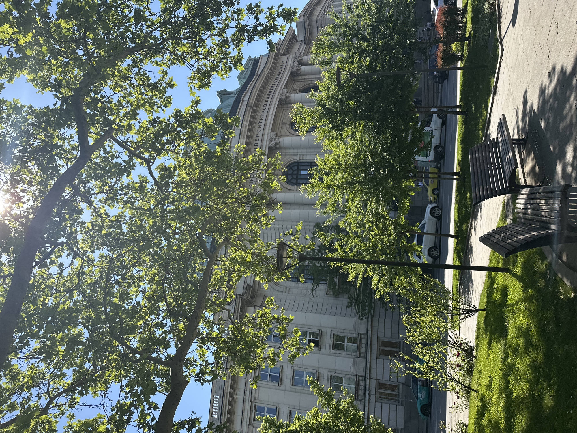 The image depicts a view of Sofia University "St. Kliment Ohridski" in Sofia, Bulgaria. The building is partially obscured by lush green trees. In the foreground, there are park benches on a grassy area adjacent to a paved sidewalk. A street with parked cars runs parallel to the sidewalk. The sky is blue and the sun is shining brightly.