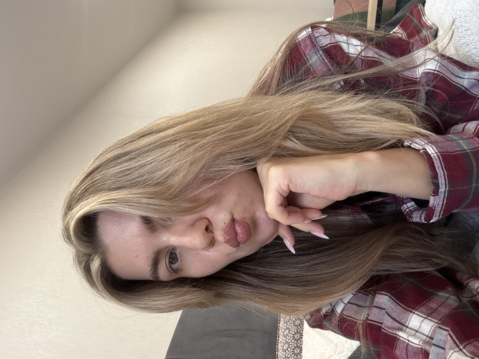 A woman with light brown hair is lying down, resting her head on her hand. She is wearing a red and gray plaid shirt. The background is a beige wall. The location is unknown.