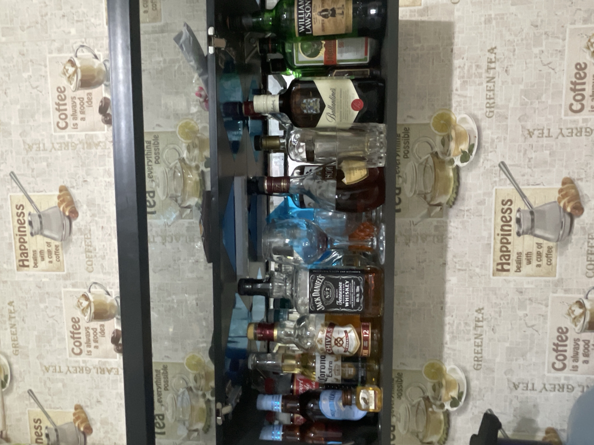 The image shows a liquor cabinet stocked with various alcoholic beverages. Brands visible include William Lawson's, Jagermeister, Ballantine's, Jack Daniel's, Chivas Regal, and Corona Extra. The cabinet is set against a backdrop of patterned wallpaper featuring tea and coffee motifs. The image suggests an indoor setting, possibly a home or bar. There's no indication of location in Sarapul, Russia, or any people or actions depicted.