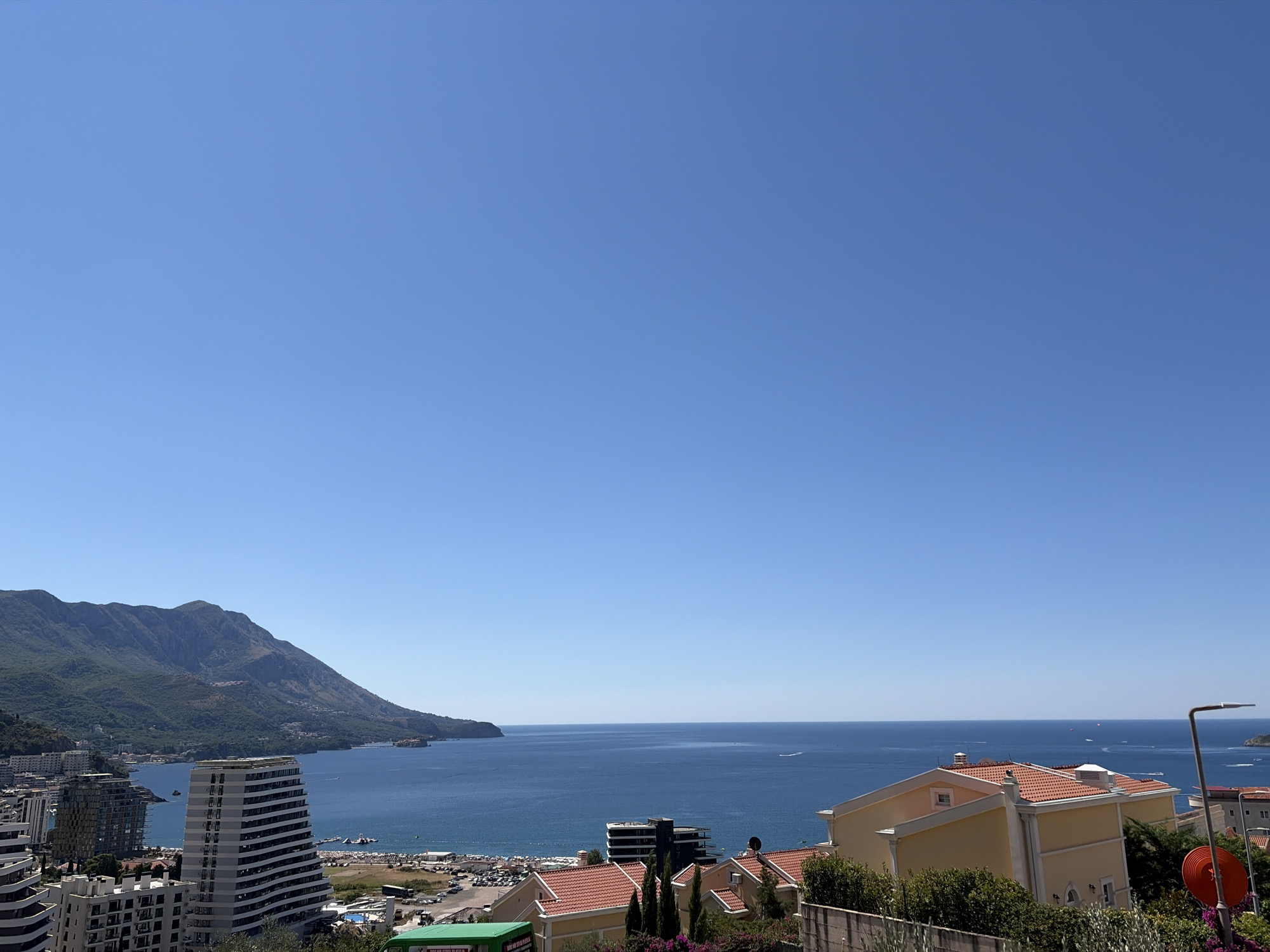 The image shows a panoramic view of the Adriatic Sea and the coastal city of Bečići, Montenegro. The sky is clear and bright blue, with no clouds visible. The sea is a deep blue, with calm waters and a few boats visible in the distance.
In the foreground, there are several buildings with red-tiled roofs. Some of these buildings appear to be residential, while others might be hotels or apartments. There are also some trees and bushes, adding greenery to the scene.
In the middle ground, the city of Bečići unfolds with a mix of modern high-rise buildings and older structures. A beach with sunbathers and umbrellas is visible, along with a parking lot filled with cars. Further out, the coastline curves, leading to a large mountain covered in lush green vegetation.
The background is dominated by the majestic mountain range that forms the backdrop of Bečići. The mountains are bathed in sunlight, with their slopes dotted with buildings and roads. The overall impression is one of a vibrant coastal town nestled against a stunning natural landscape. The scene appears to be captured during daytime, likely in the late morning or early afternoon, given the strong sunlight and clear visibility.
