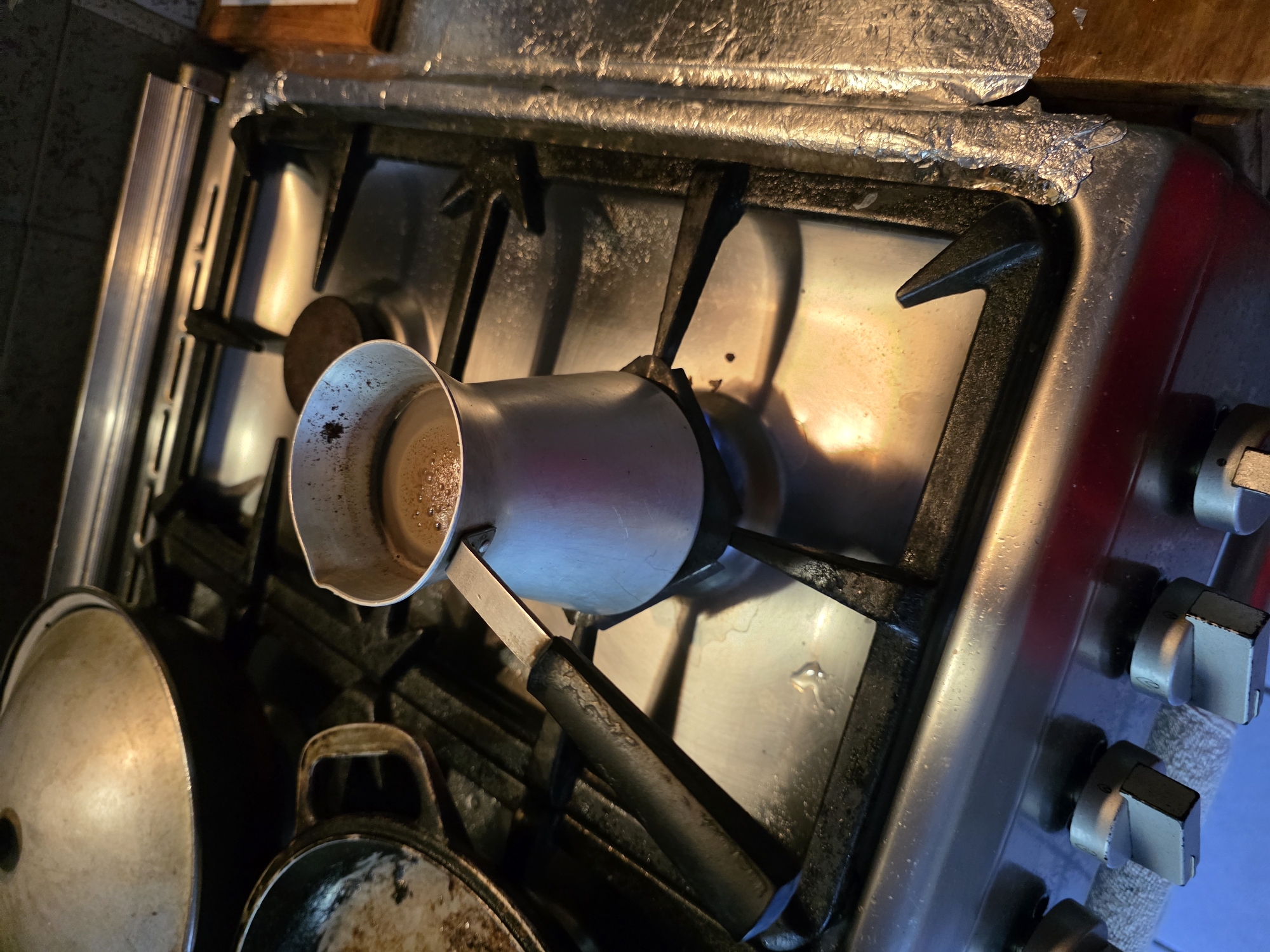 This media file depicts a close-up, overhead view of a gas stove in a kitchen setting. The primary subject is a moka pot, a stovetop coffee maker, positioned on one of the burners. The pot is made of metal and appears to contain some liquid with a foamy surface, suggesting that coffee is either being brewed or has recently been brewed.
Other objects visible include other pots and pans on the stove, some of which appear to be cast iron, and a metal lid. The stove itself is a standard gas model with metal grates and control knobs. The surface of the stove shows signs of use, with some splatters and a generally unpolished appearance. There's also a piece of aluminum foil haphazardly placed on the backsplash above the burners, possibly to catch grease or protect the surface.
The lighting in the scene is warm and somewhat dim, with highlights reflecting off the metal surfaces of the stove and cookware. This suggests it might be indoors during the evening or in a room with limited natural light. The overall impression is of a functional, lived-in kitchen. There are no people visible in the image.
The location context provided is Tuapse, Russia, but there are no specific visual cues within the image that definitively confirm this location. The scene is consistent with a kitchen found in many parts of the world.
There is no visible text in the image.