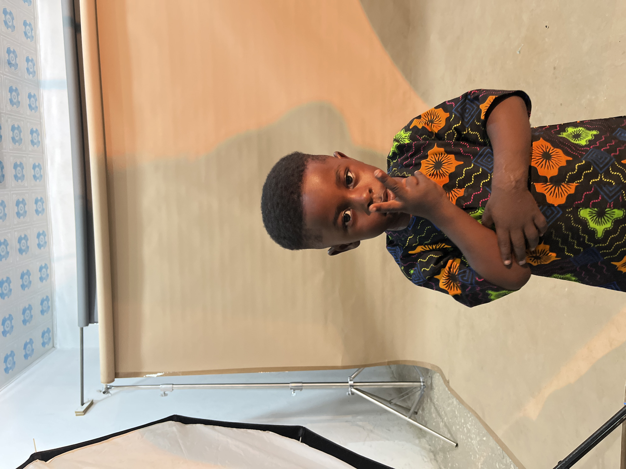 A young Black child with short dark hair is posing in a studio. The child is wearing a colorful shirt with an orange and green floral pattern against a black background. Their right hand is raised to their face, with fingers touching their lips. The child's gaze is directed at the camera with a curious expression.
The setting appears to be a photography studio, indicated by a backdrop that transitions from a light beige color to a darker beige, and a photographic light diffuser in the foreground. To the left, there is a wall with a repeating blue and white tiled pattern. The lighting suggests it is daytime, possibly indoors with artificial studio lights. The child is standing, and the overall scene is well-lit. No text is visible in the image. The overall mood is playful and engaging.