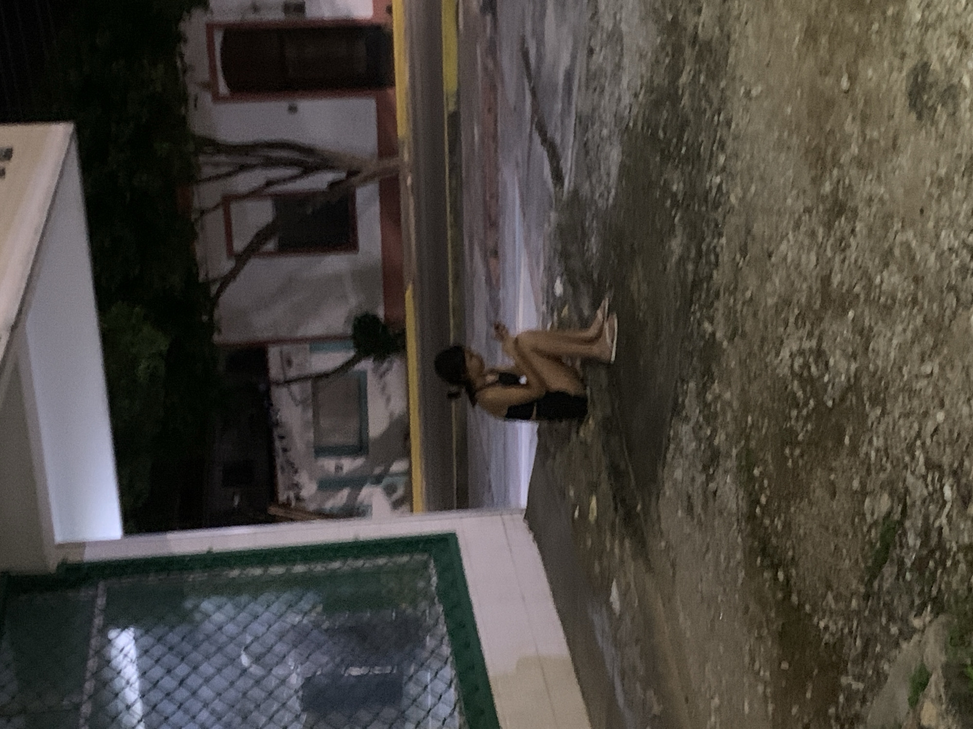 In Santa Ana, Venezuela, a young woman with dark hair tied back sits on a curb at night, looking down at her phone. She wears a dark tank top and shorts, with light-colored sandals on her feet. The pavement around her is dirty and uneven, with some debris scattered on it. Behind her, a building with white walls and red trim is visible, along with a tree. The street in front of the building has a yellow line painted along its edge. To the left of the scene, a green-framed window with a mesh covering is part of a building's facade. The overall lighting suggests an outdoor urban environment after dark.