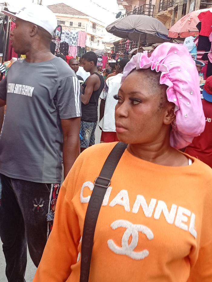 The image captures a busy street market scene in Fegge, Nigeria, during daylight. In the foreground, a dark-skinned woman faces left, her profile partially visible. She wears an orange long-sleeved top with "CHANNEL" and the Chanel logo embroidered in white, and a shiny pink, possibly satin, bonnet adorned with small, clear embellishments. A dark strap from a bag crosses her upper torso. To her left, a dark-skinned man is visible from the waist up, facing right. He wears a white baseball cap, a grey short-sleeved t-shirt with white stripes on the sleeves and "FUTURE SPORTSWEAR" printed on the chest, and dark grey sweatpants featuring a red and black pattern on the side and a black ninja-like character logo on the thigh.
In the mid-ground and background, multiple other individuals are present, including a man in a black tank top and patterned jeans, and another bald man in a white shirt. Merchandise, consisting primarily of clothing items such as t-shirts and sleepwear, is displayed on racks and hangers along the street. Some garments feature cartoon characters. Multi-story buildings line the thoroughfare, and several market umbrellas, including a prominent dark grey one, are visible, providing shade over stalls. The overall lighting suggests natural daylight.